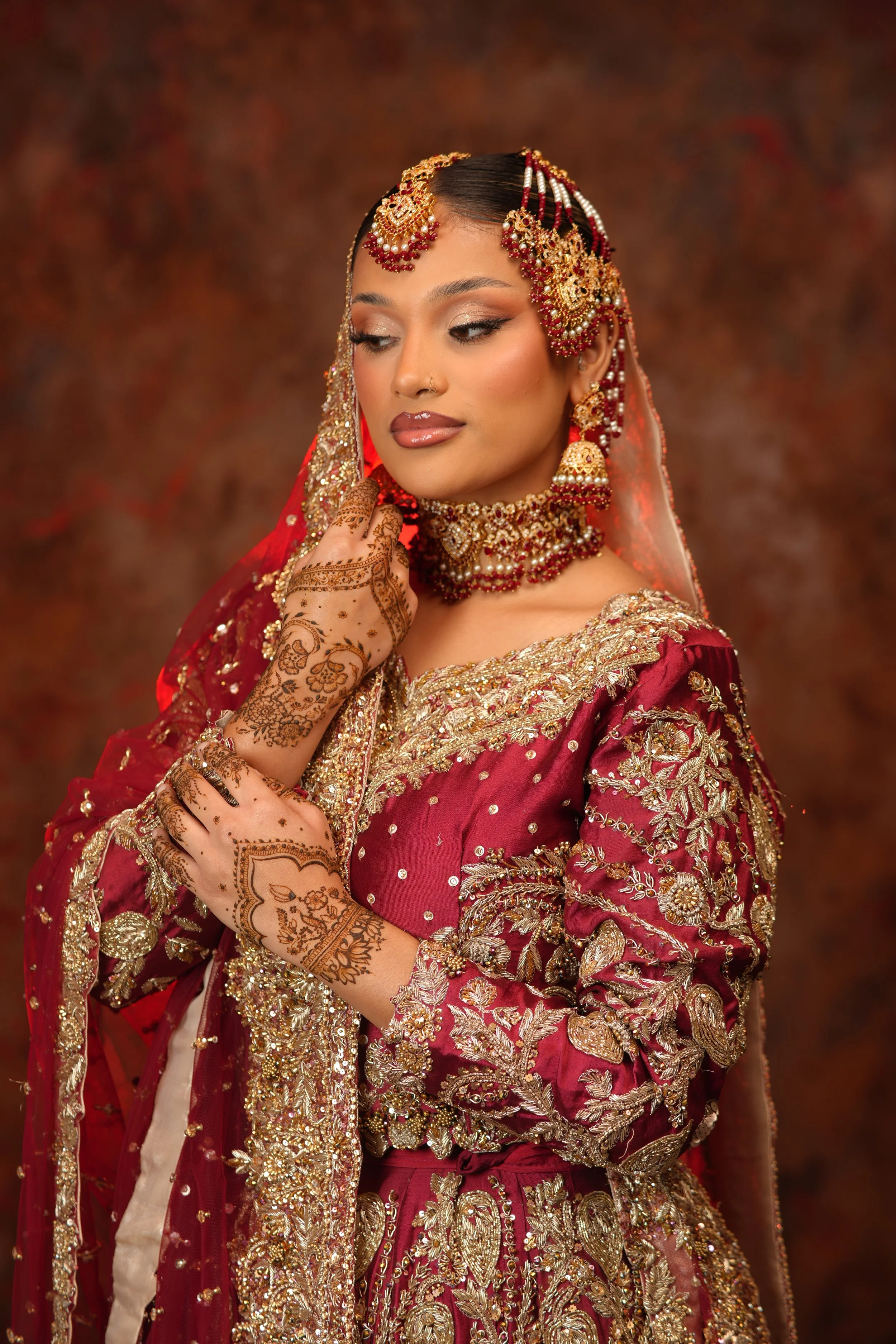 Indian bride in red lehenga and red dupatta draped over her head, with soft smokey eyes, fluffy lashes, glowing skin, and red lipstick. Bridal henna decorates the hand resting on her shoulder.