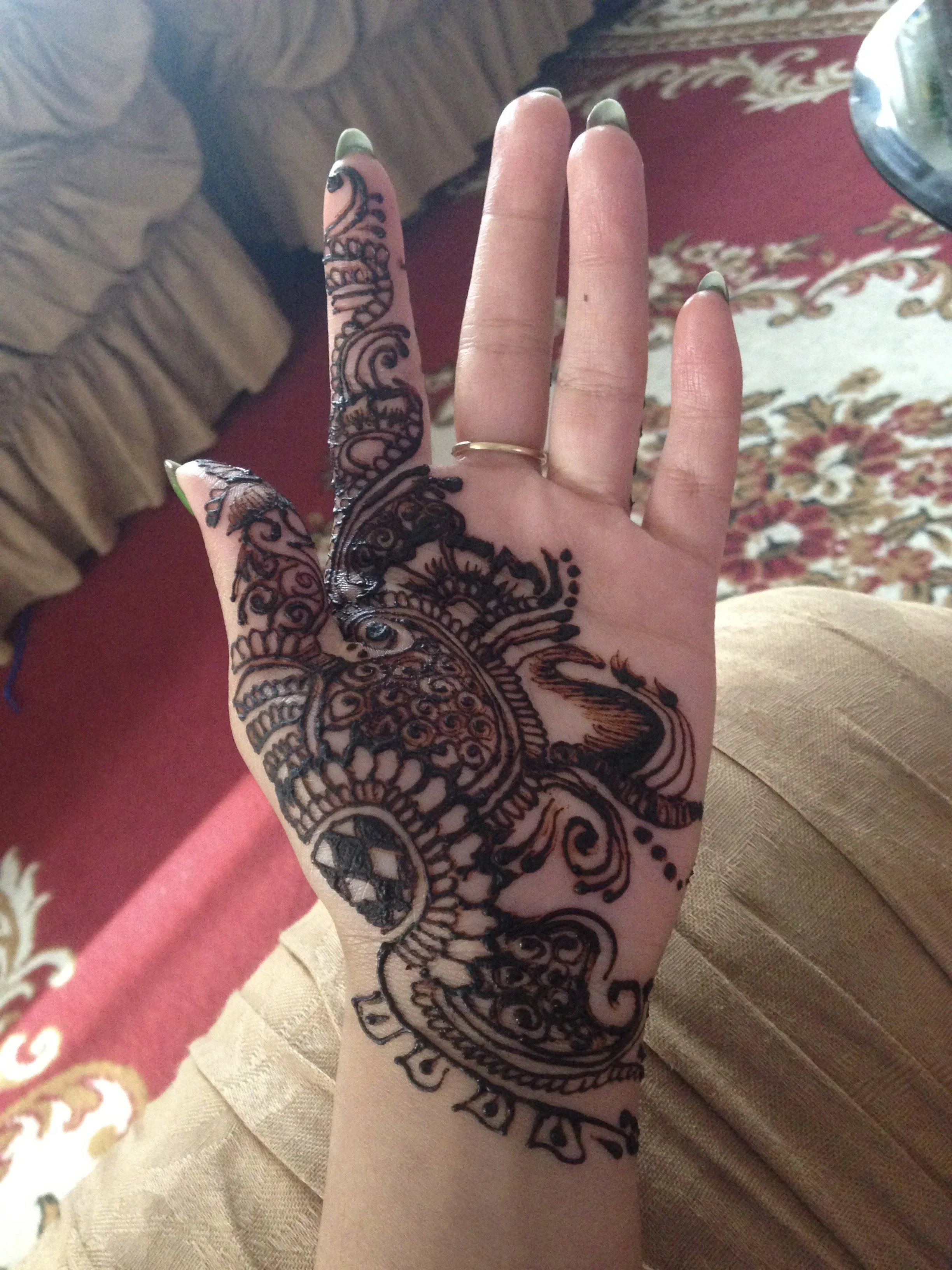 A hand with intricate black henna designs on the back, featuring paisleys, floral motifs, and swirling patterns, with long green-painted nails and a gold ring on the middle finger.