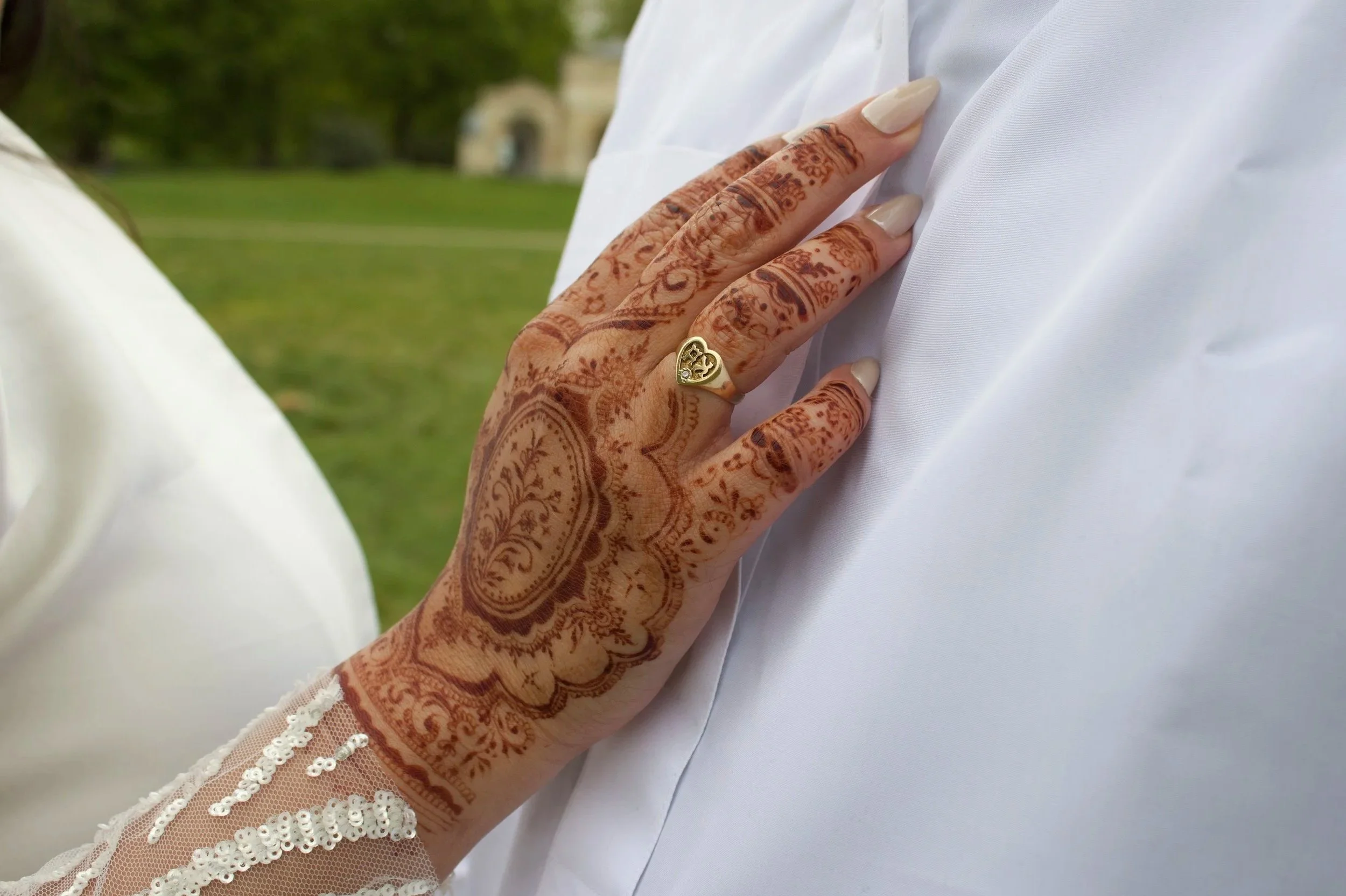 Close-up of a woman's hand with henna designs touching a man's white shirt. The woman is wearing a gold ring with a heart-shaped design, and her fingernails are painted. The background shows a green outdoor setting.