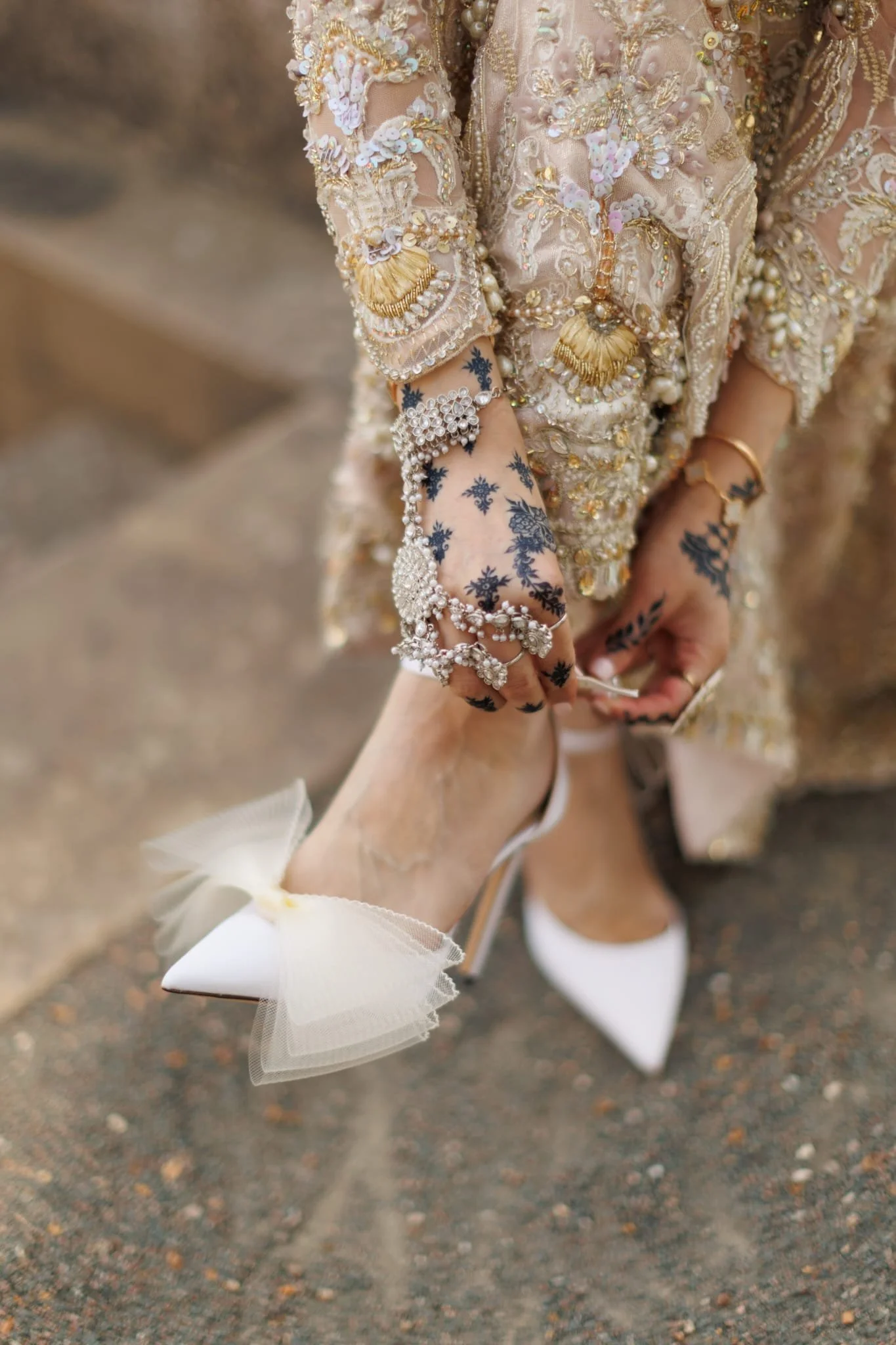 Close-up of a woman wearing a detailed, embroidered dress with gold and pearl embellishments, adjusting her white high-heeled shoe with a large decorative bow, intricate black henna designs on her hand, and wearing jewelry including rings, bracelets, and an anklet.