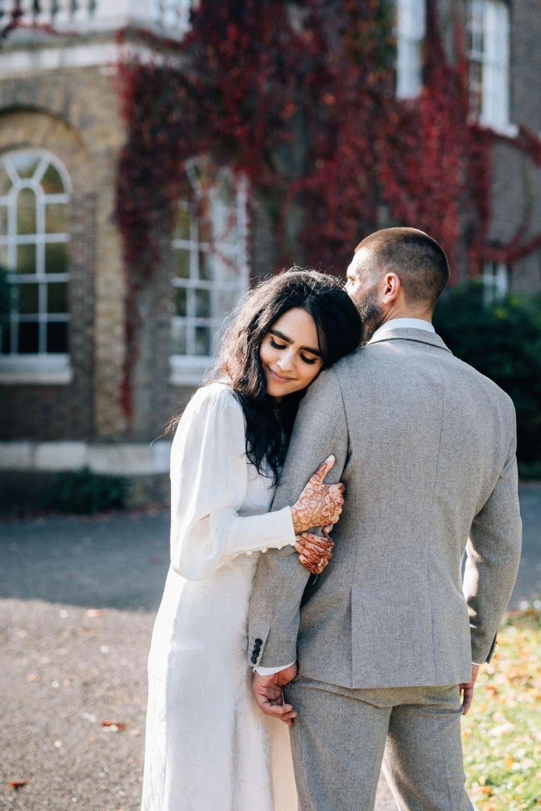 A woman with henna tattoos on her hands leaning her head against a man's shoulder outdoors on a sunny day, with a background of a brick building covered in red ivy.