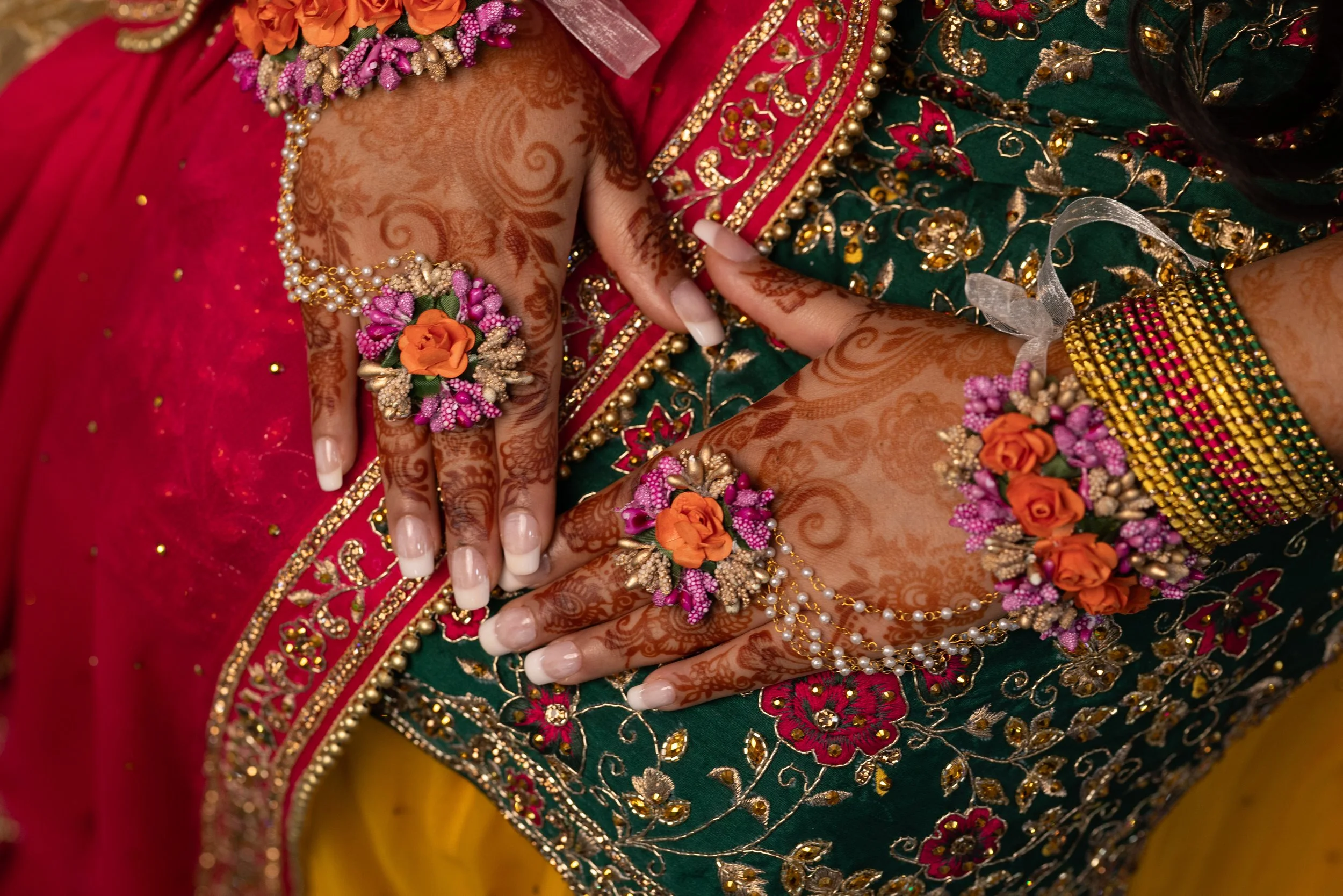 Close-up of hands decorated with henna, adorned with flower jewelry, rings, and bracelets, against vibrant traditional colorful embroidered clothing.