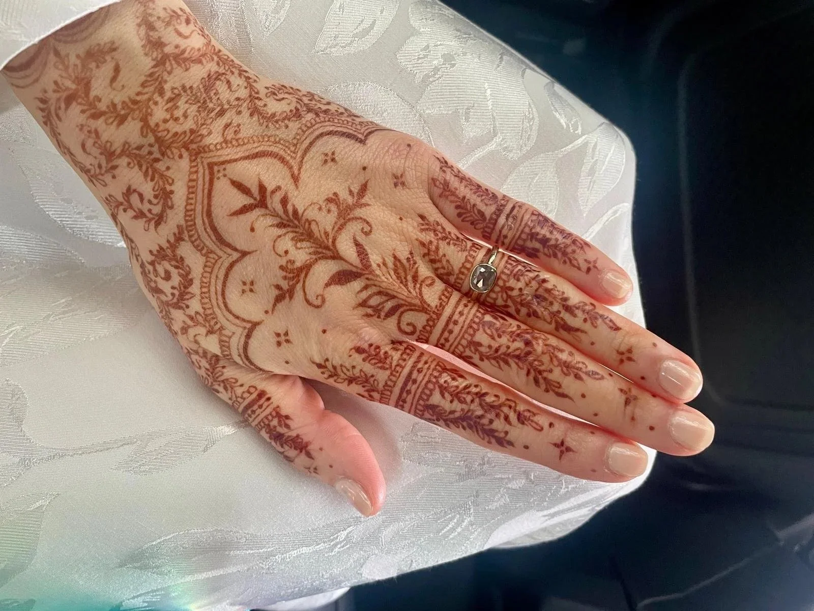 Close-up of a hand with intricate henna designs on the fingers and back of the hand, featuring floral and geometric patterns, wearing a silver ring, resting on white fabric with a subtle leaf pattern.