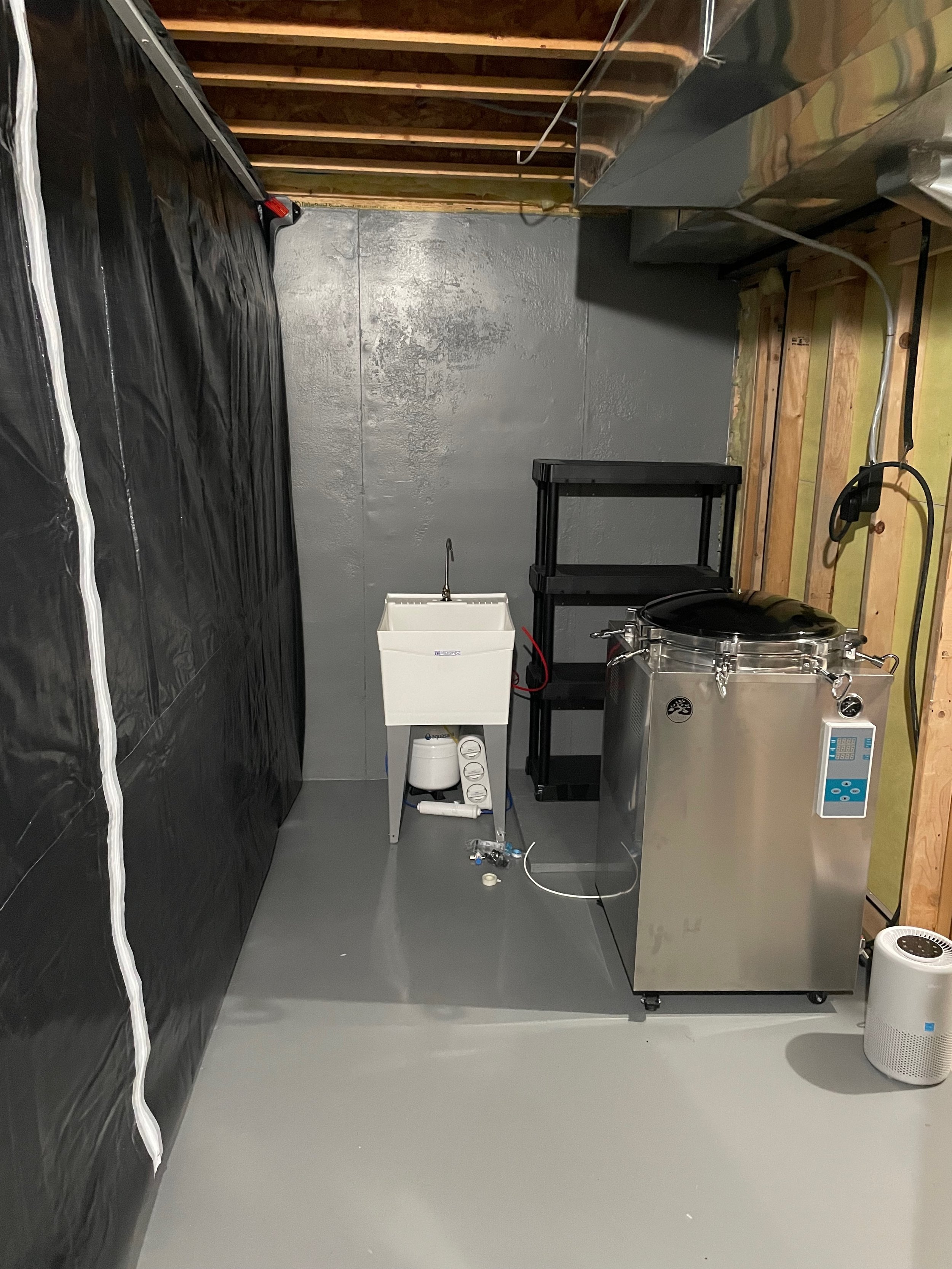 Basement area with a reverse osmosis water filtration system, large stainless steel autoclave, black storage shelves, and a plastic curtain.