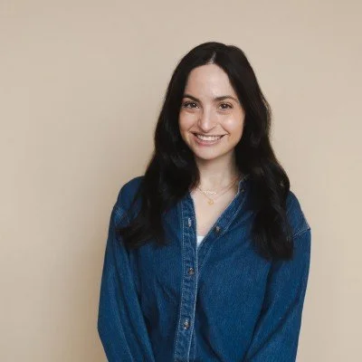 A young woman with long dark hair smiling, wearing a blue denim shirt, standing against a neutral beige background.
