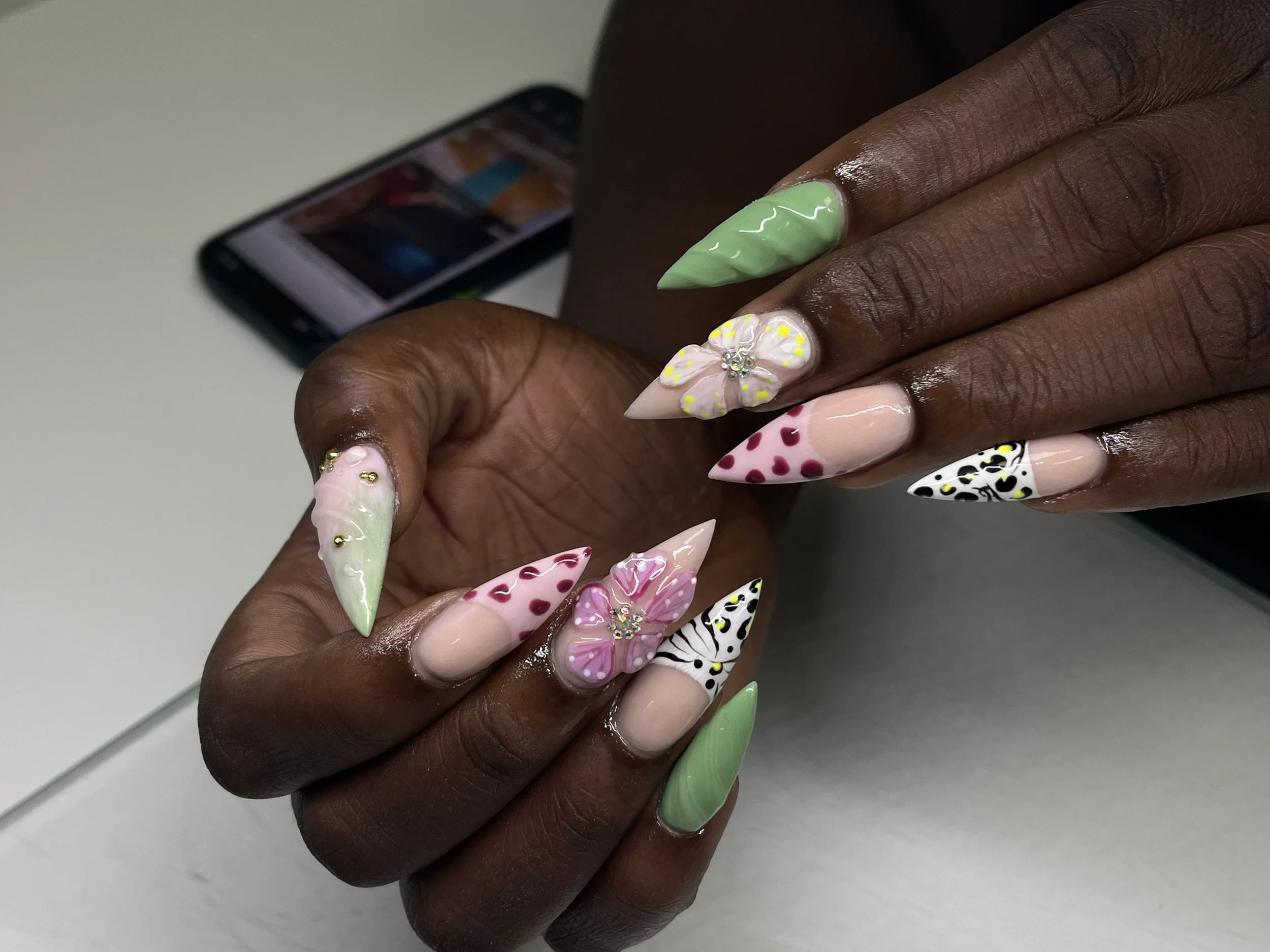 Close-up of a person's hand with long, decorated nails featuring floral and animal print designs in pastel and bright colors, with a smartphone in the background.