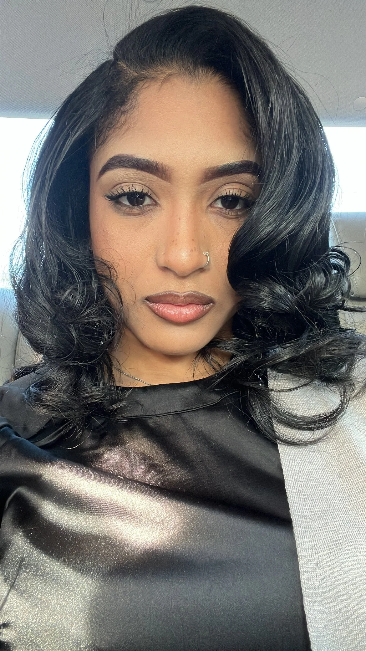 Close-up of a woman with black, styled hair, wearing makeup, a nose ring, and a shiny black top, sitting inside a vehicle.