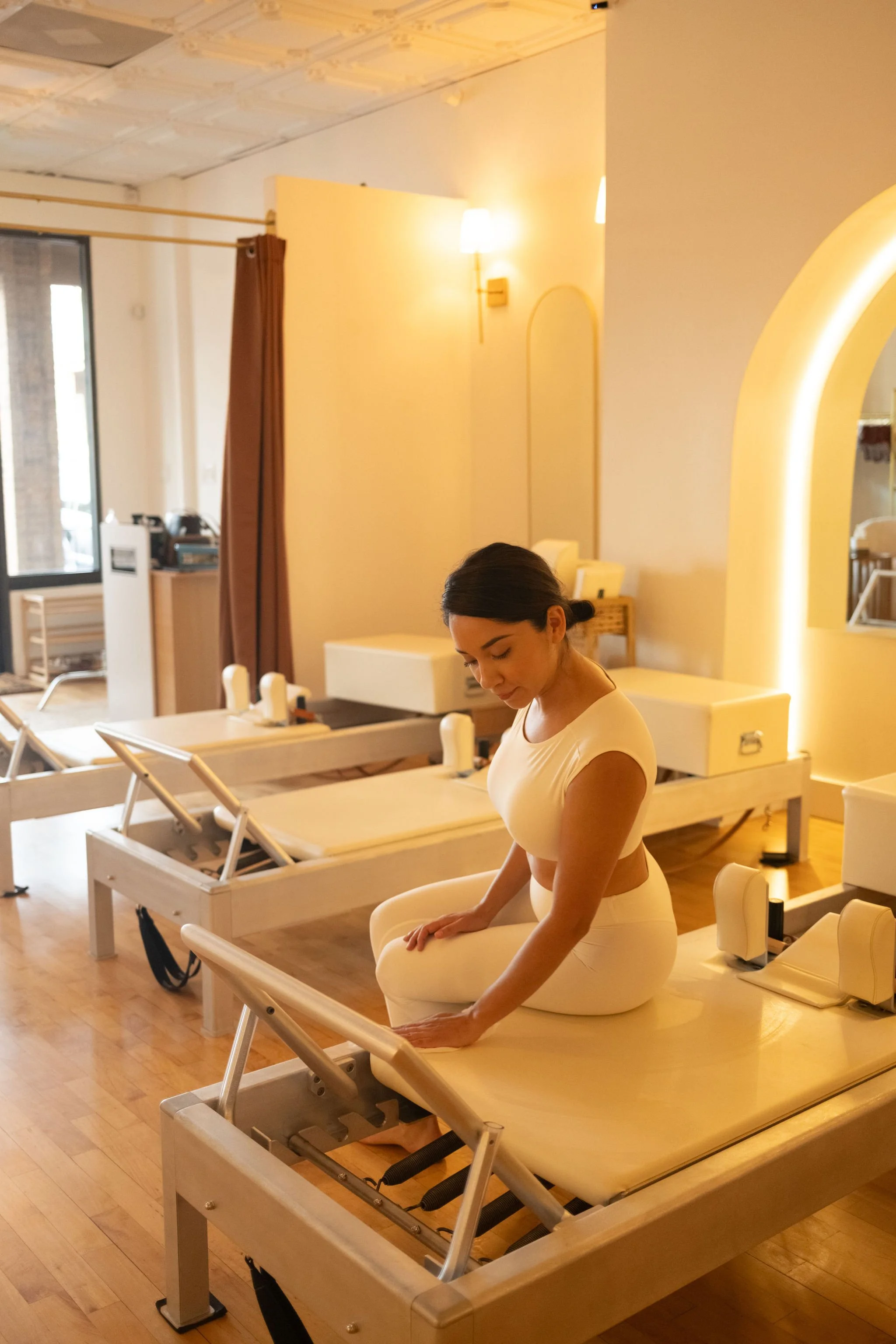 A woman in a white workout outfit is seated on a reformer in a well-lit room with wooden floors and white walls. She appears to be cleaning the Pilates apparatus.