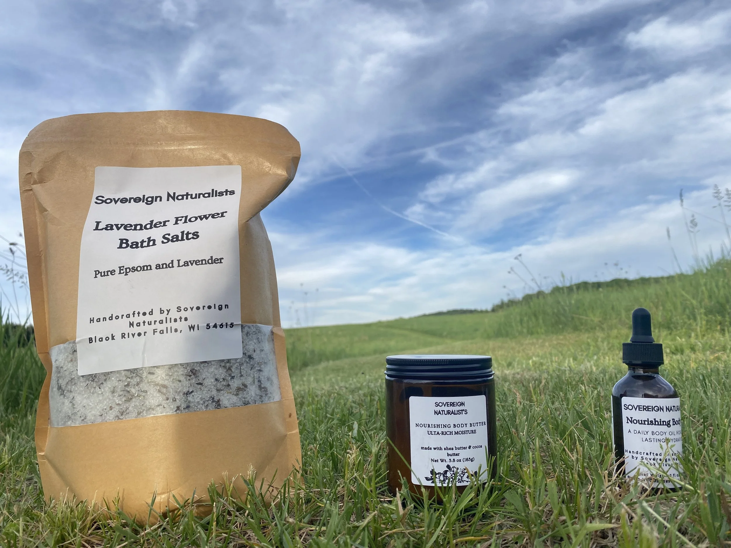 Packaged lavender bath salts, natural skincare products, and body butter bottles placed on grass outdoors with a blue sky and clouds in the background.