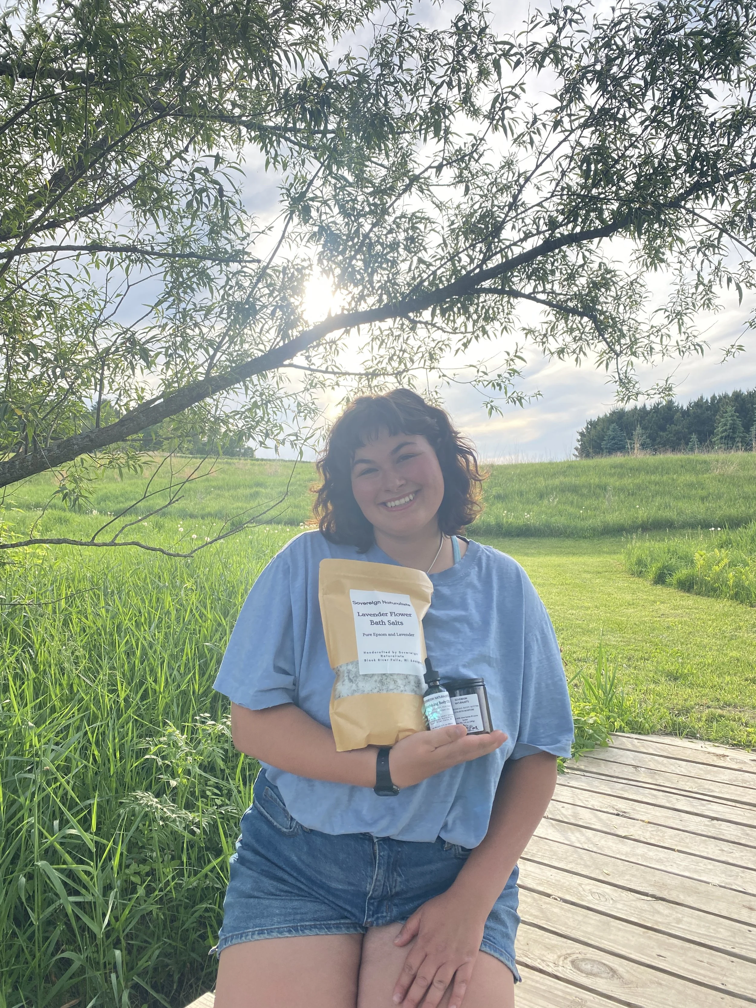 A smiling woman with curly hair wearing a blue t-shirt and denim shorts, holding a brown paper bag and a small bottle, standing on a wooden walkway outdoors with green grass, trees, and cloudy sky in the background.
