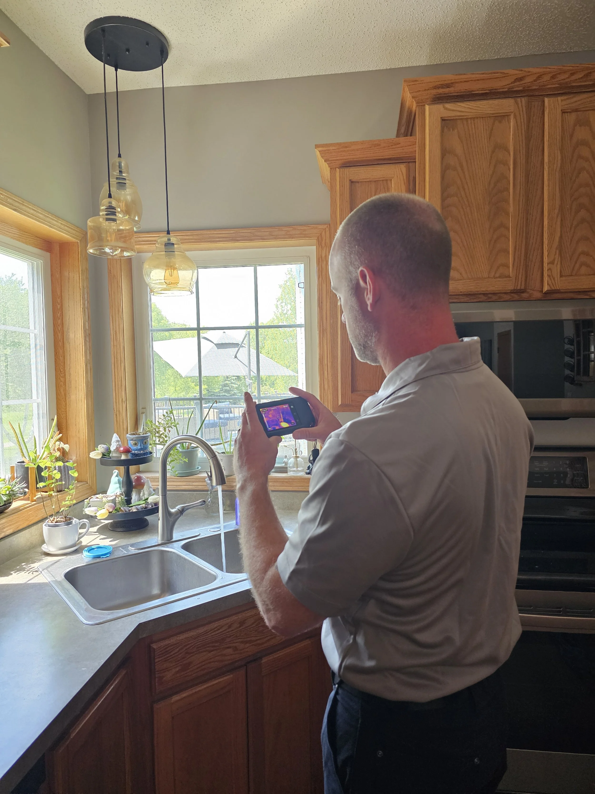 Trailblazer home inspector using a handheld thermal camera to scan the area around a kitchen sink for hidden water leaks or moisture intrusion.