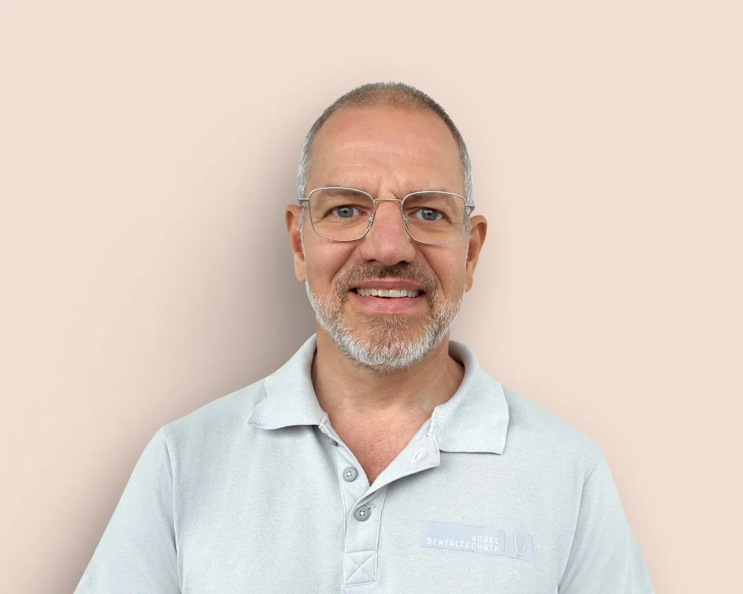 Experienced dental technician and managing director with glasses, gray hair and beard in a polo shirt with the "Vogel Dentaltechnik" logo against a light background - stands for trust, expertise and successful cooperation with Noevu.