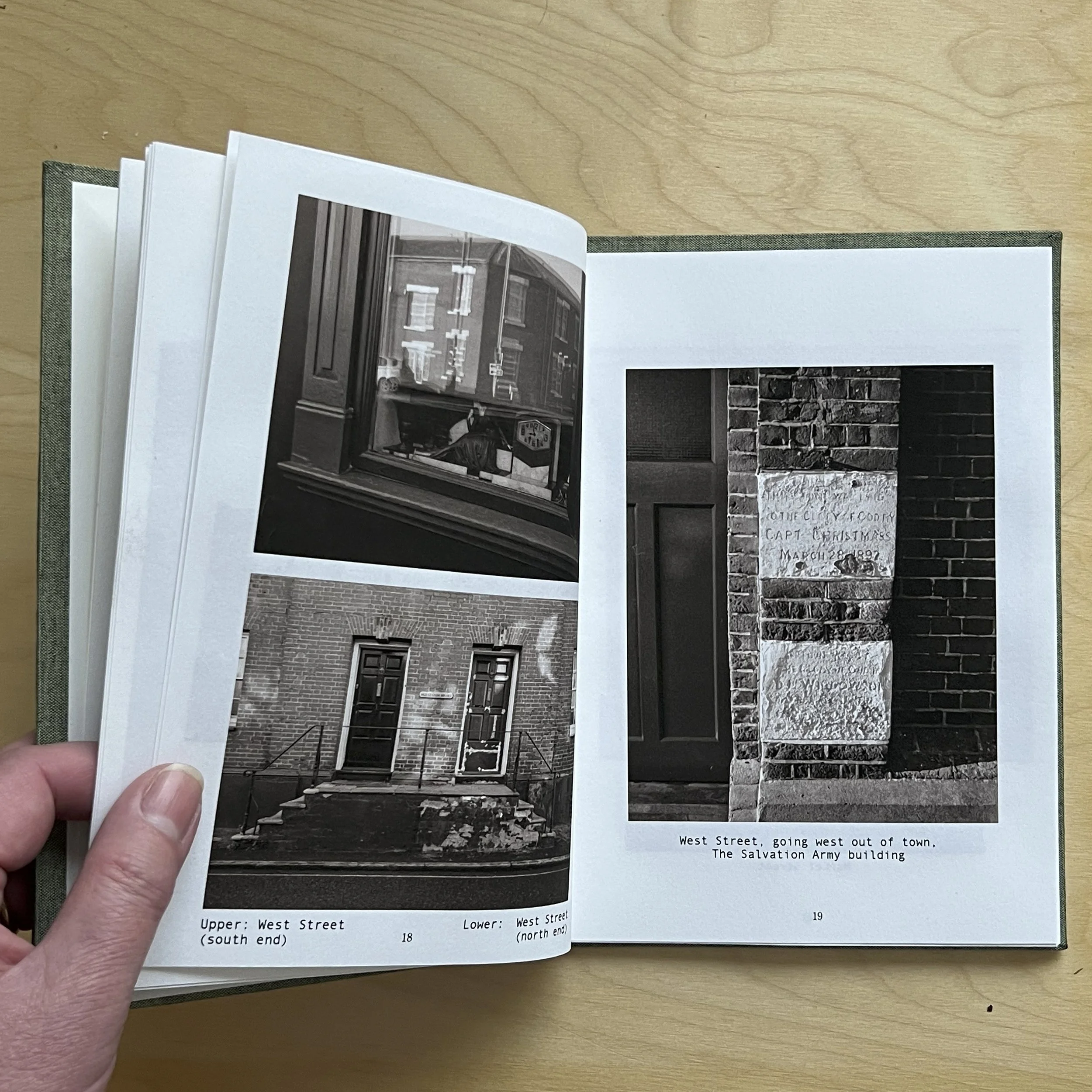 Open photo book displaying black and white photos of West Street buildings, including a storefront window, building steps, and a brick wall with a plaque reading 'City of Cory, Capt. Christmas, March 28, 1897'.
