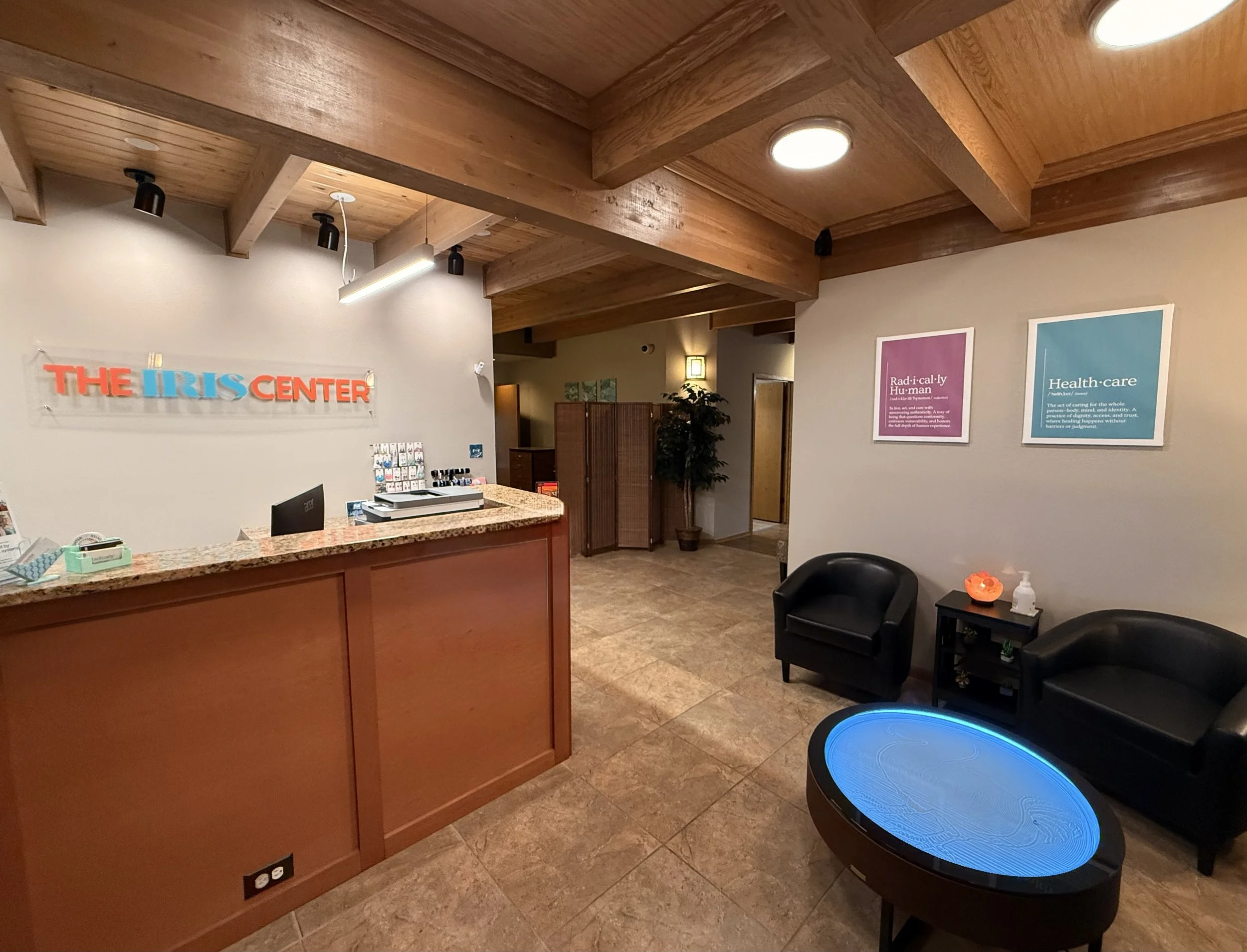 Reception area of The IRIS Center with a wooden desk, signs, and seating. A wall sign says 'The IRIS Center' in colorful letters. The seating includes two brown leather chairs, and the walls are decorated with framed informational posters. The space has wood ceiling beams and a tiled floor.