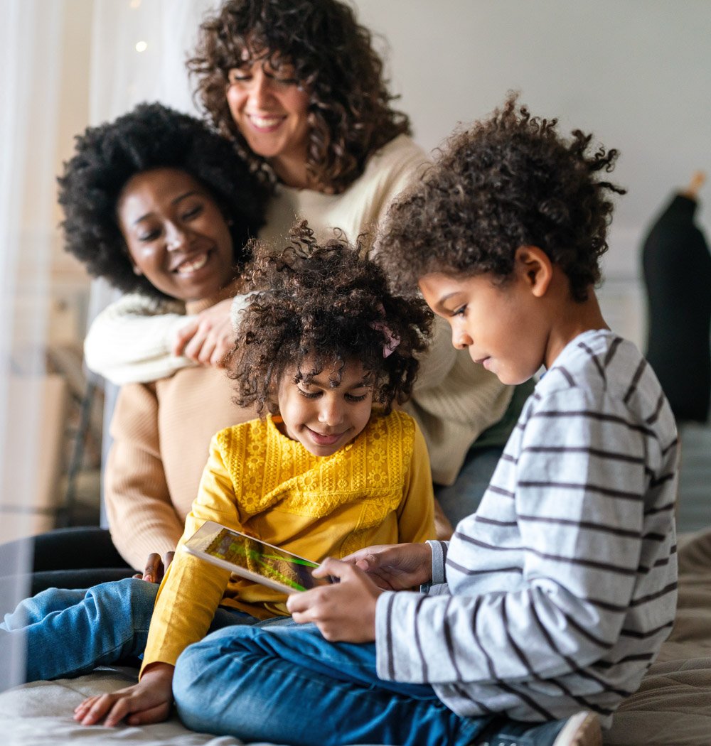 A woman and three children with curly hair are sitting closely indoors, with the woman smiling and looking at the children. One boy is showing the girl a tablet device, and she is leaning in to look at it.