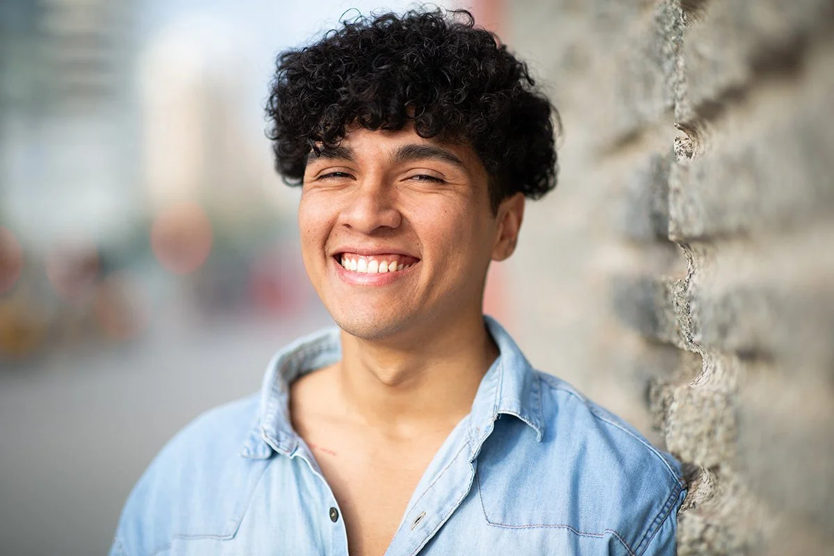 Smiling young man with curly dark hair standing against a brick wall.