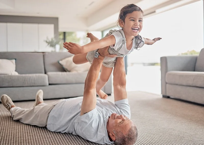 A man lying on the carpet playing with a young girl, who is flying through the air with her arms outstretched in a living room with large windows and modern furniture.