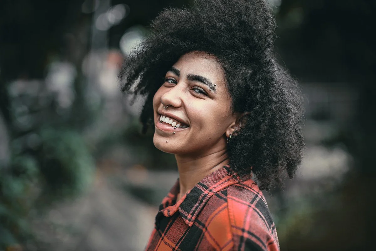 Person with curly hair smiling, wearing a red plaid shirt, and having facial piercings.