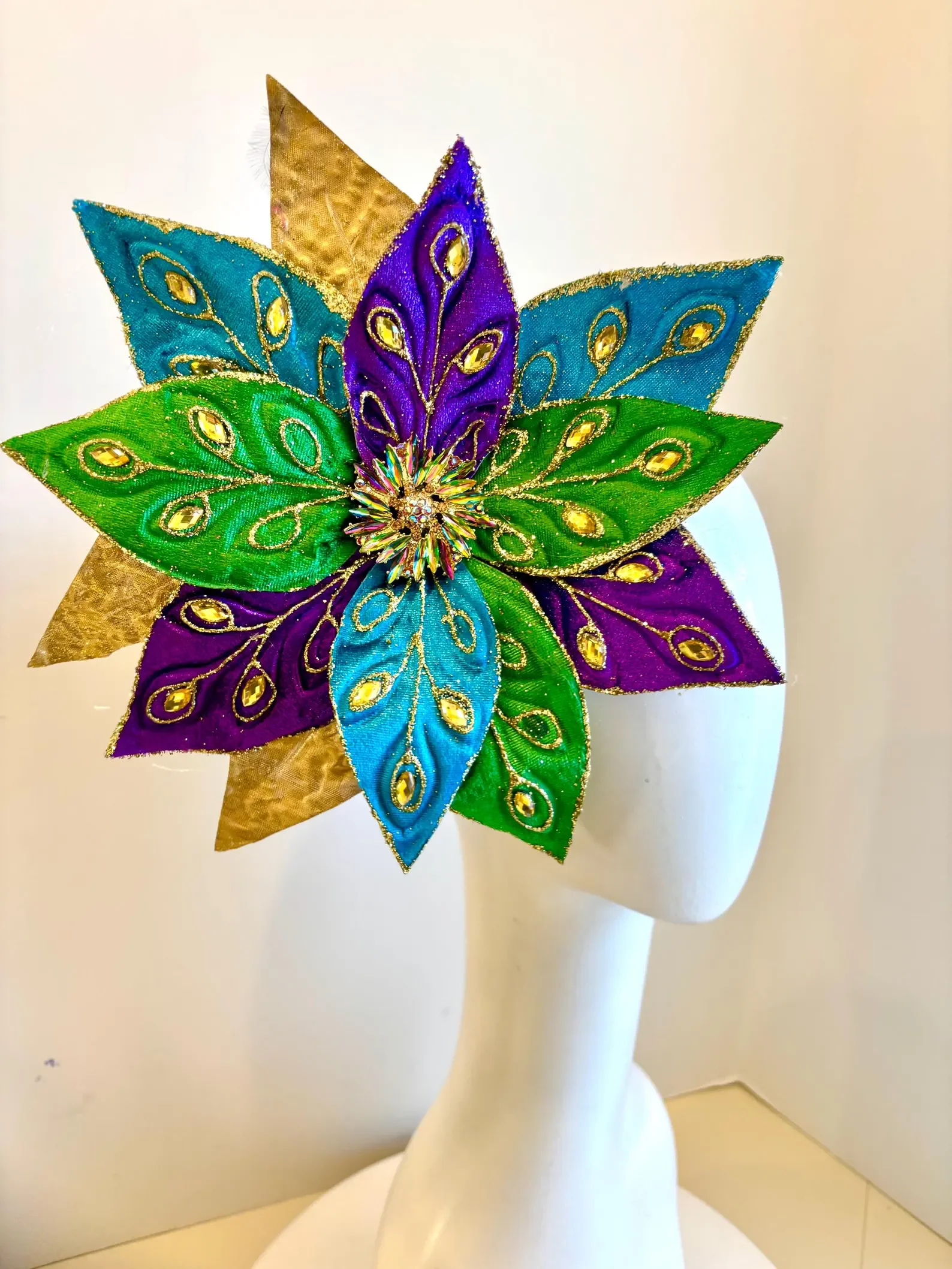 Colorful decorative headpiece with large, layered petals in gold, purple, blue, and green, adorned with gold accents and beaded details, displayed on a white mannequin head.