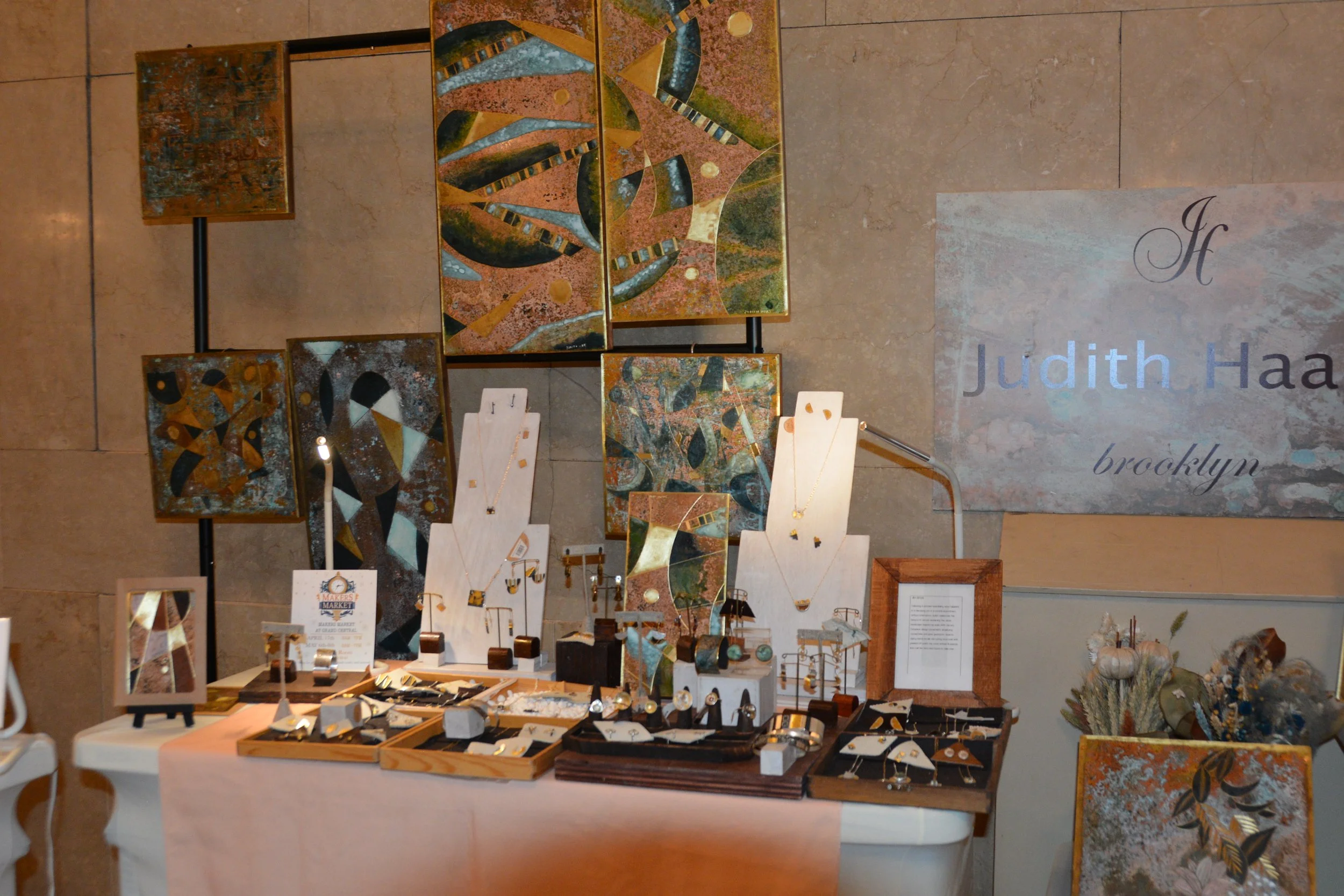 Jewelry display booth featuring necklaces, earrings, and rings, with abstract artwork in the background and a sign reading 'Judith Haa Brooklyn'.