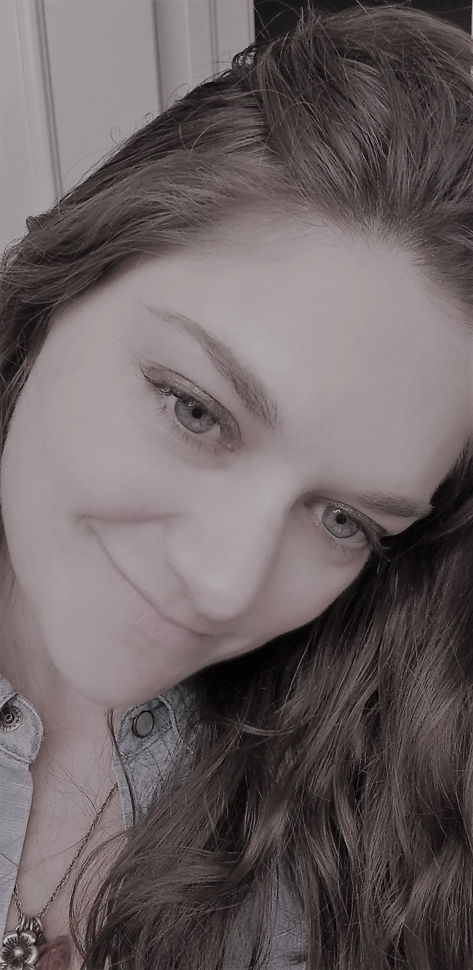 Close-up of a young woman with long wavy hair, light skin, light-colored eyes, smiling, wearing a necklace and a button-up shirt.