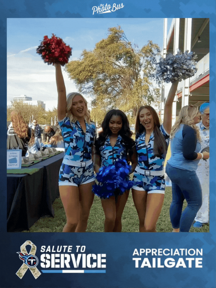 Three cheerleaders in blue camouflage uniforms celebrating at a tailgate event, holding pom-poms, with a Tennessee Titans logo and salute to service banner in the foreground. Taken by the Roamer Photo booth.