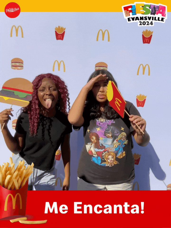 Two young women holding McDonald's fries and burgers, posing playfully at a themed event in front of a backdrop with McDonald's logos and cartoon fast food images. The event is called Fiesta Evansville 2024. Taken with a branded social booth.