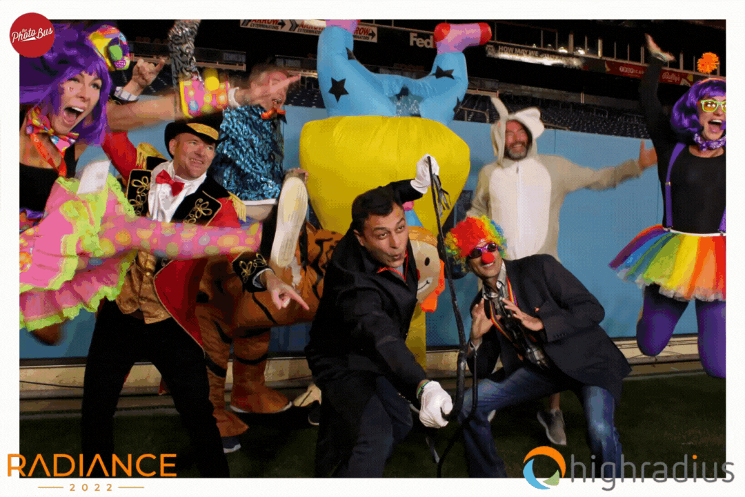 A group of people dressed in colorful clown and carnival costumes, some wearing wigs and vibrant accessories, posing for an array photo booth that creates a 3D image.