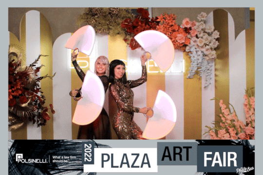 Two women in elegant dresses posing for an array photo booth at the Plaza Art Fair with decorative floral arrangements and art installations in the background.
