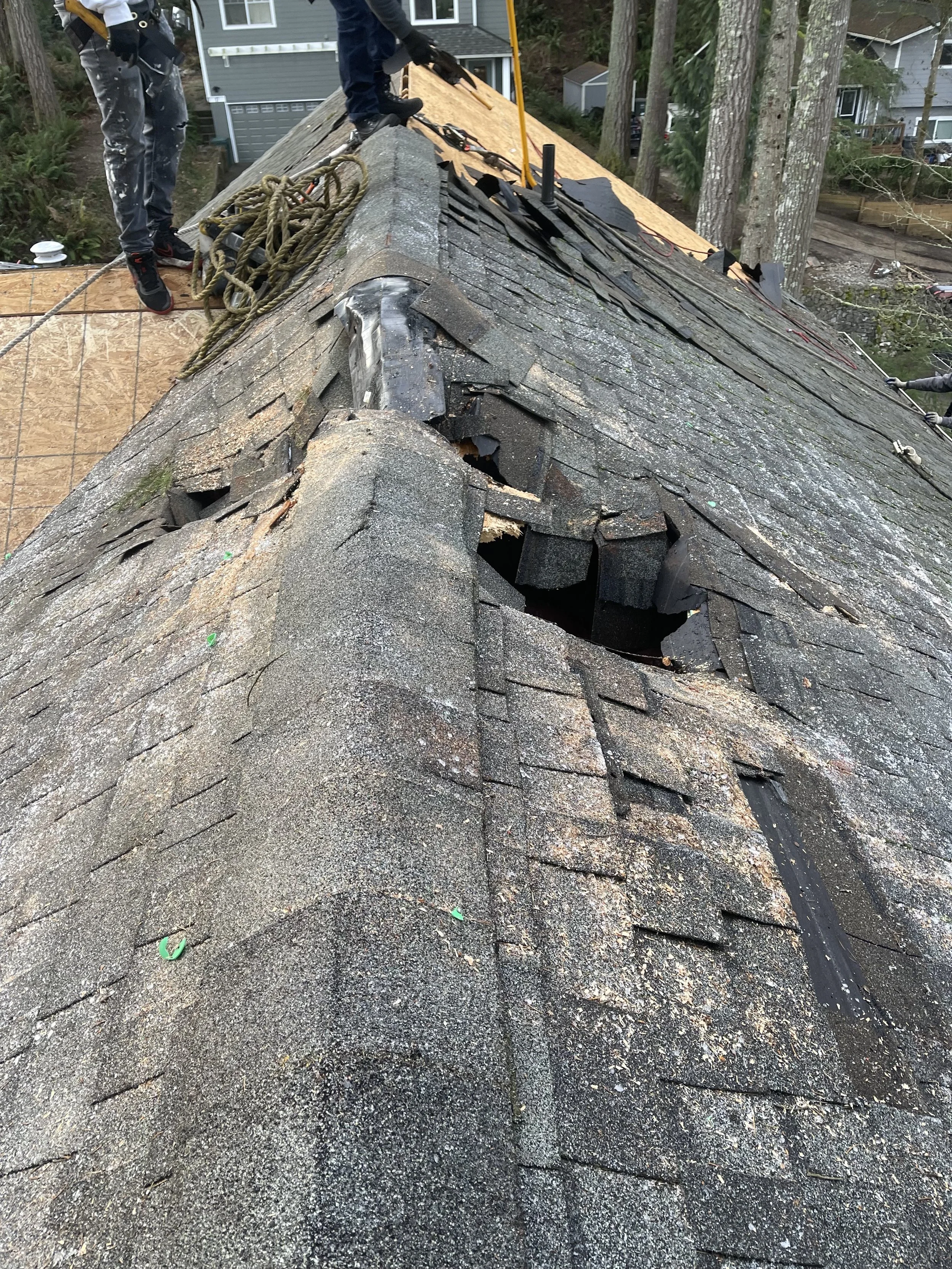 Repair workers fixing a damaged roof with missing and broken shingles, using safety ropes in a residential area.