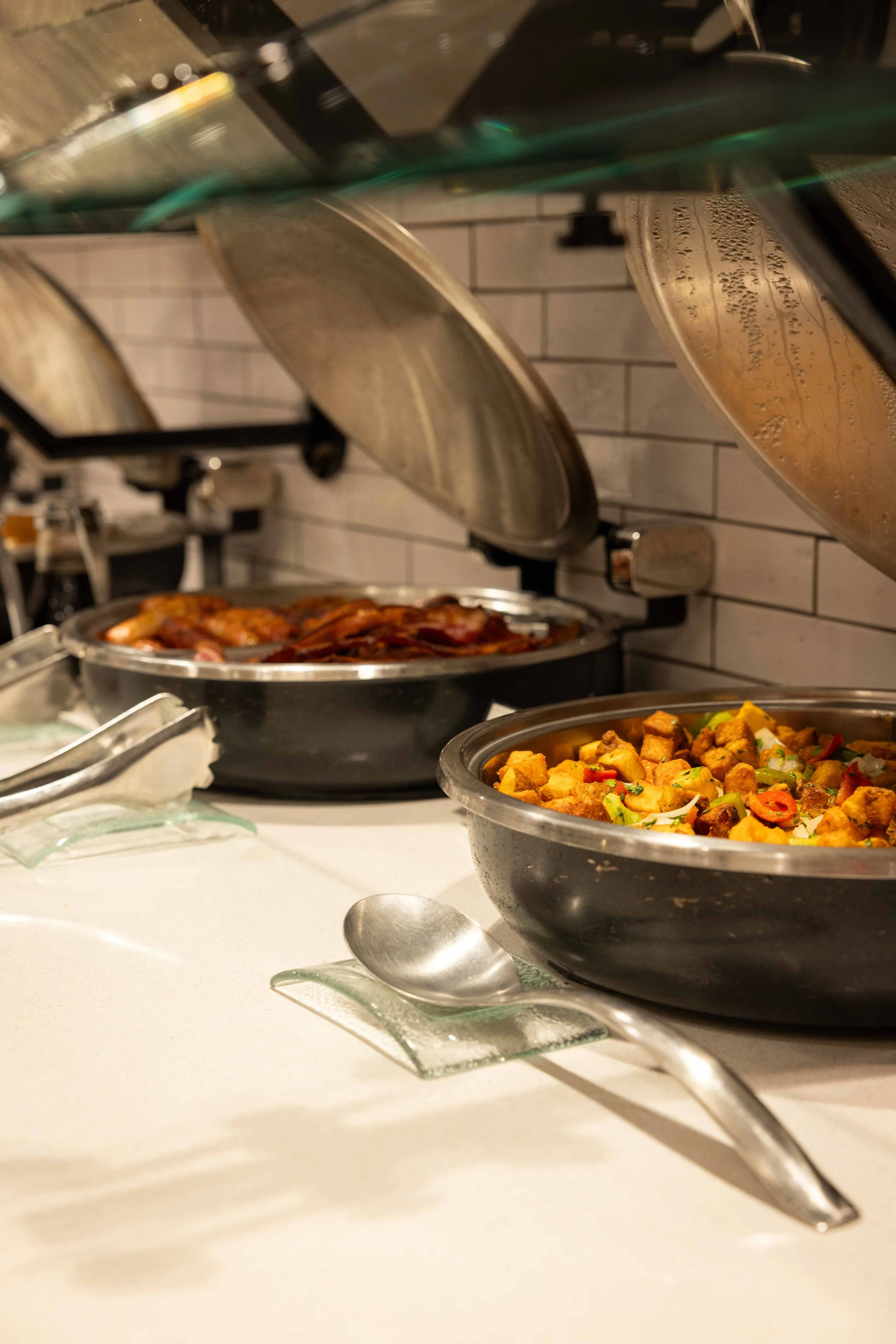Buffet with stainless steel chafing dishes filled with cooked food, including roasted meats and vegetables, in a restaurant or catering setting with a white tiled wall background.