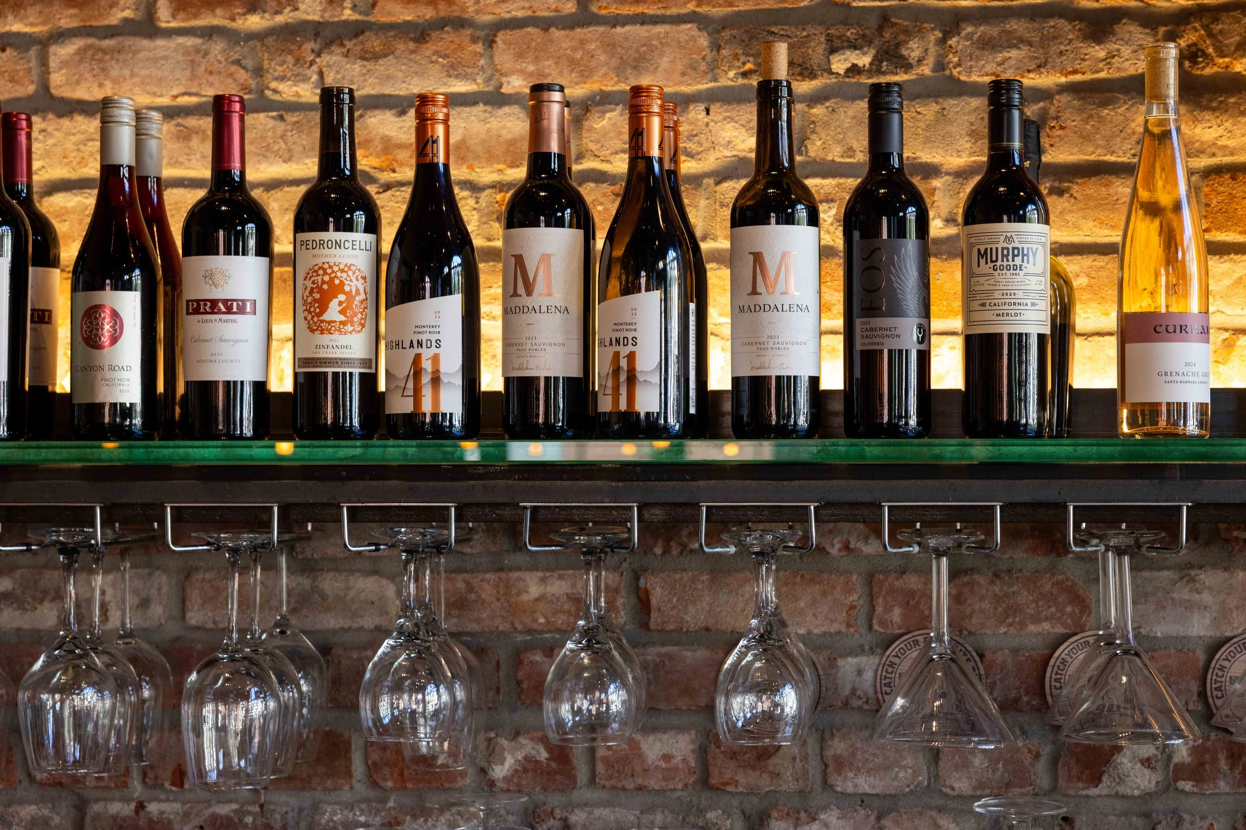 A row of wine bottles on a glass shelf above upside-down wine glasses, against a brick wall background.