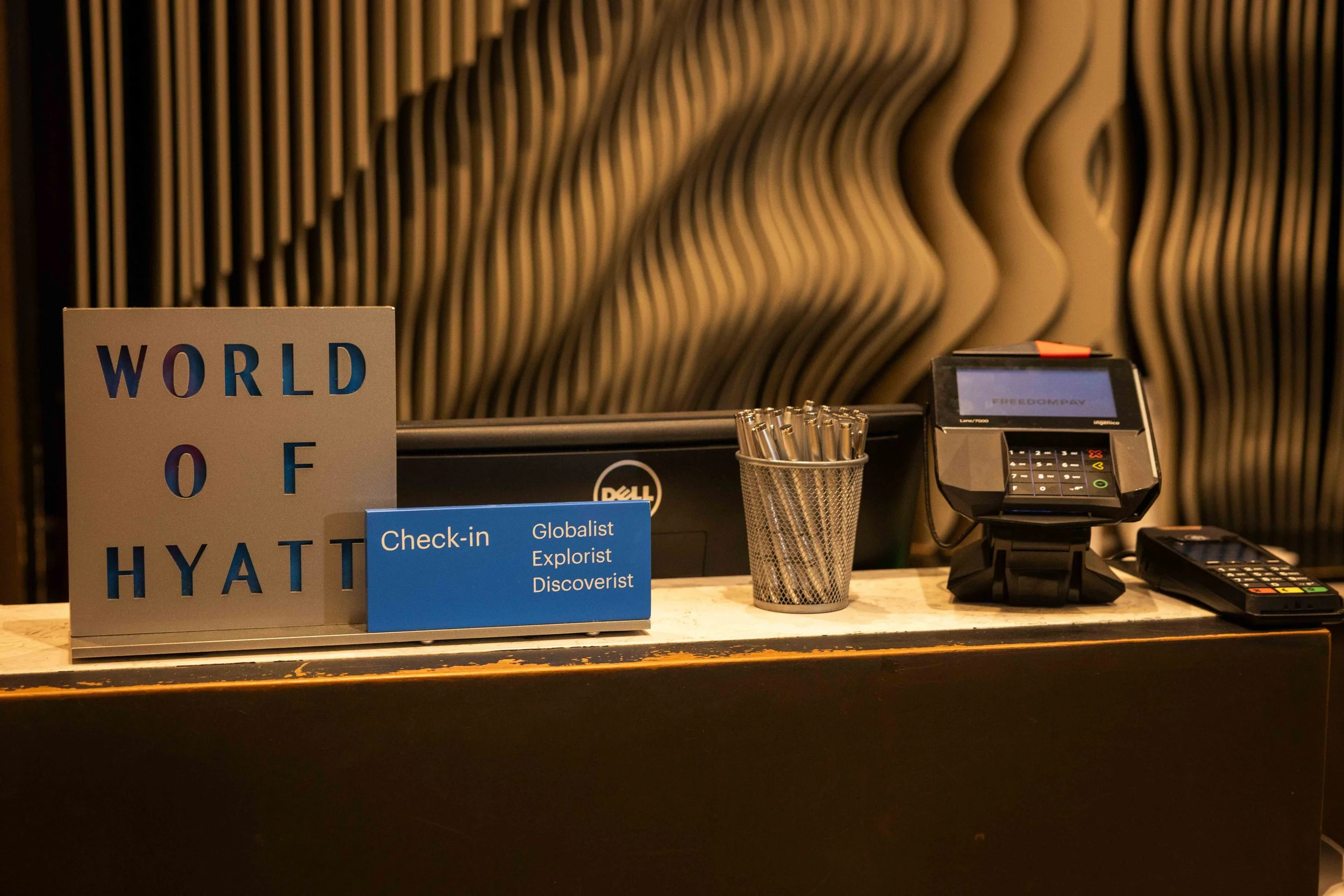 Hotel check-in counter with a sign reading 'World of Hyatt,' a small blue sign listing 'Check-in,' 'Globalist,' 'Explorer,' and 'Discoverist,' a pen holder with pens, a payment terminal, and a computer monitor in front of a textured, wavy wall.