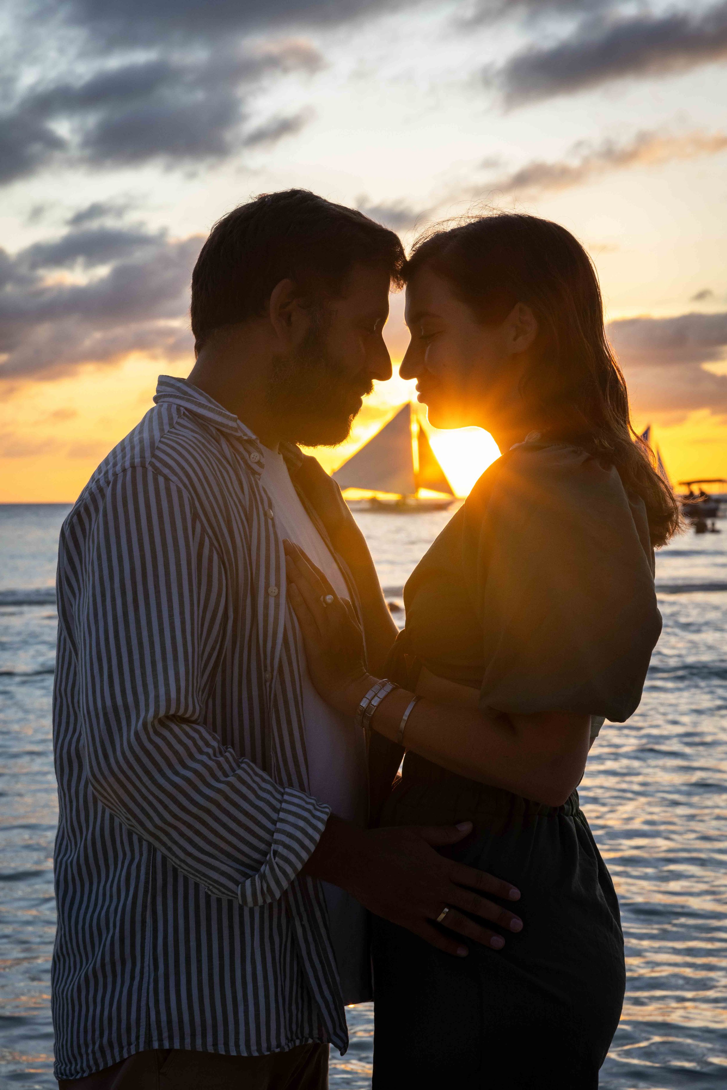 Romantic couple embracing at sunset near the ocean with a sailboat in the background. Boracay beach lifestyle family photoshoot by Anna Meshcheri