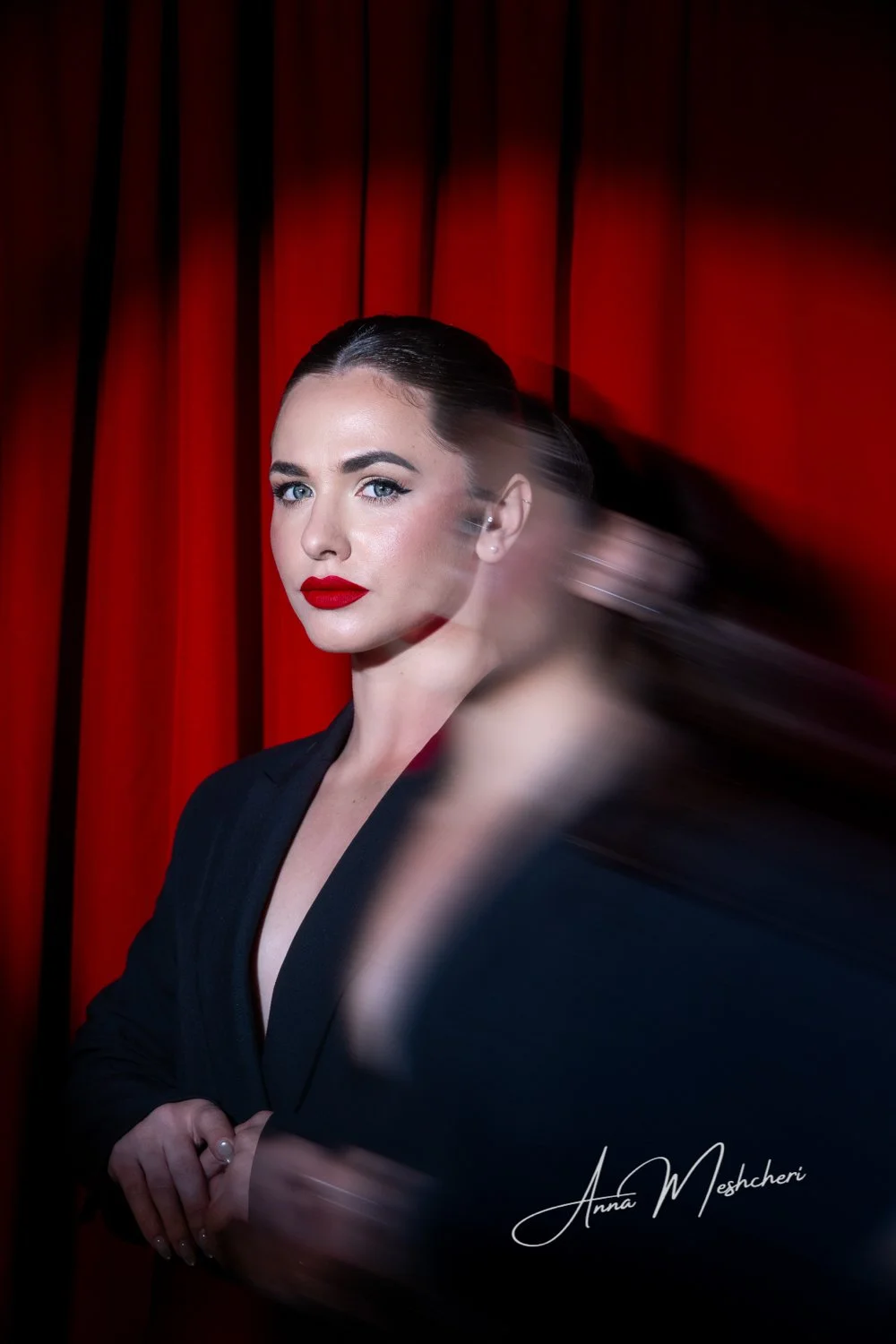 Creative fashion portrait with motion blur effect featuring a model in a black blazer and red lipstick photographed against a dramatic red studio drape backdrop.