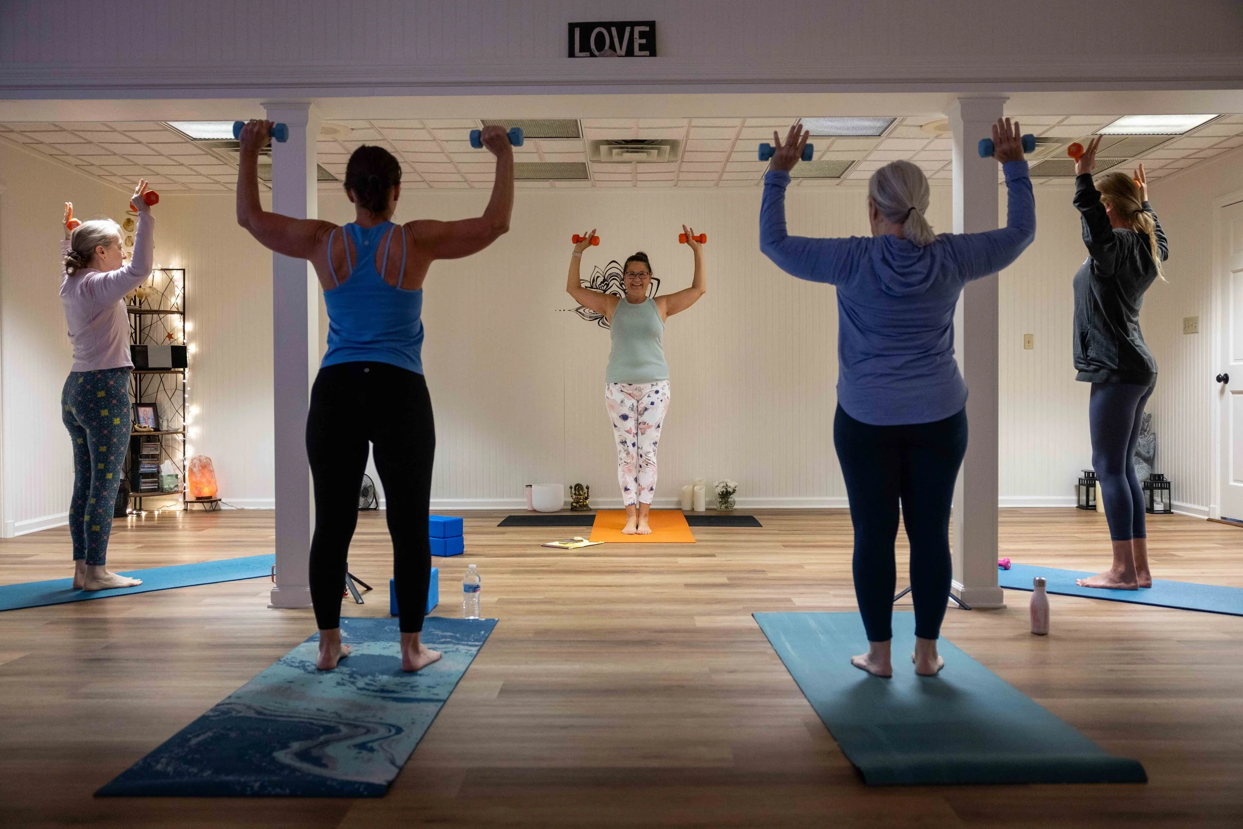A group of women practicing yoga with weights in a studio, led by an instructor at the front. They are on yoga mats, raising their arms holding small dumbbells during a pose.
