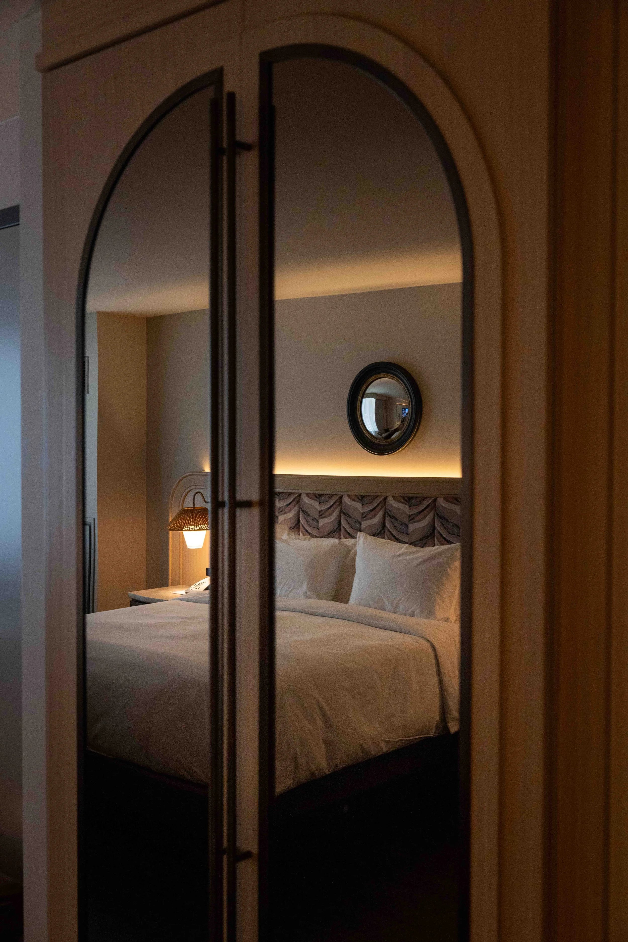 A hotel room visible through a mirror reflection, featuring a bed with white linens, pillows, a patterned headboard, a bedside lamp, and a small round mirror on the wall.