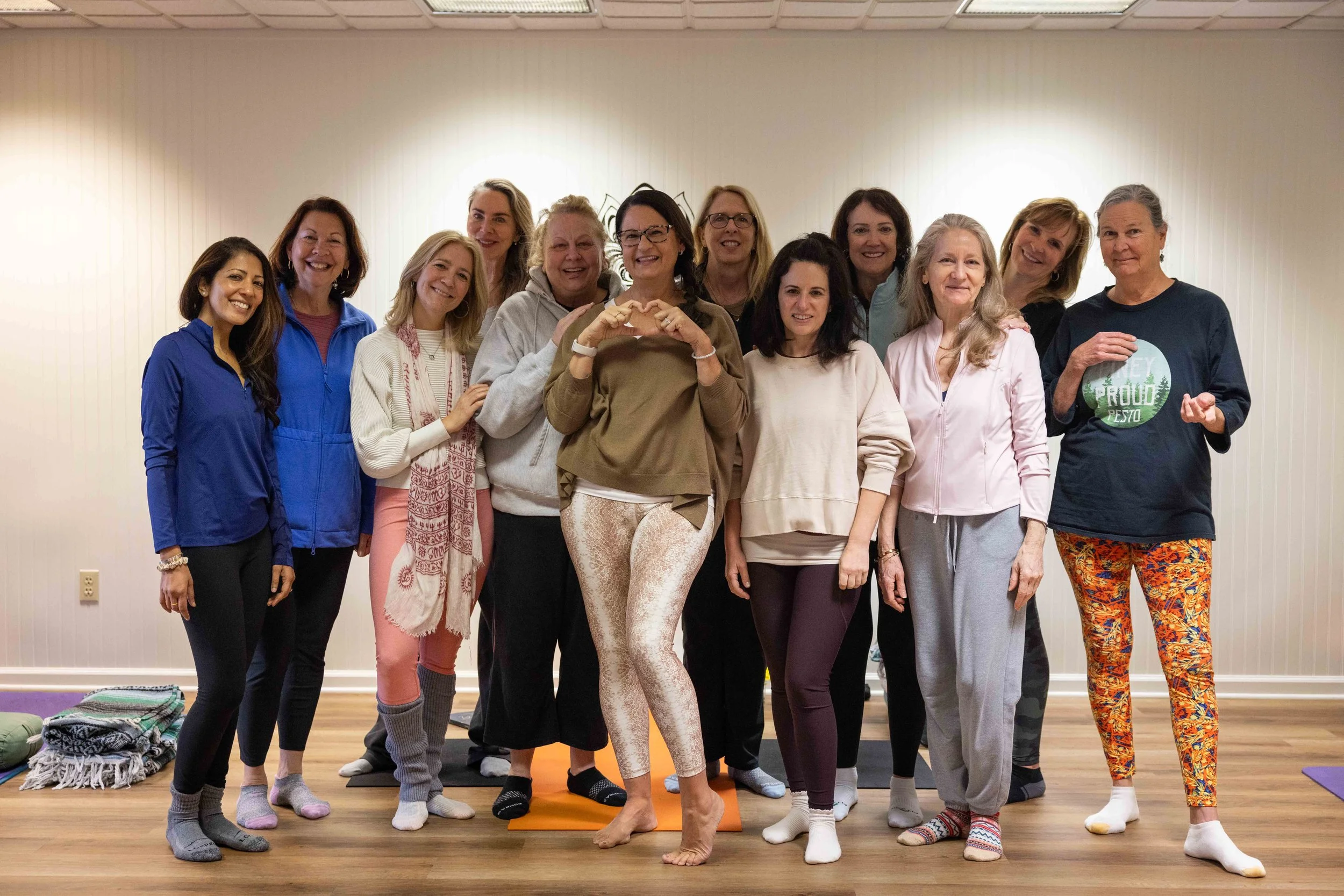 A group of women gathered in a yoga studio, posing for a photo with some smiling and others making hand gestures. They are dressed casually, and the environment suggests a relaxed, friendly atmosphere.