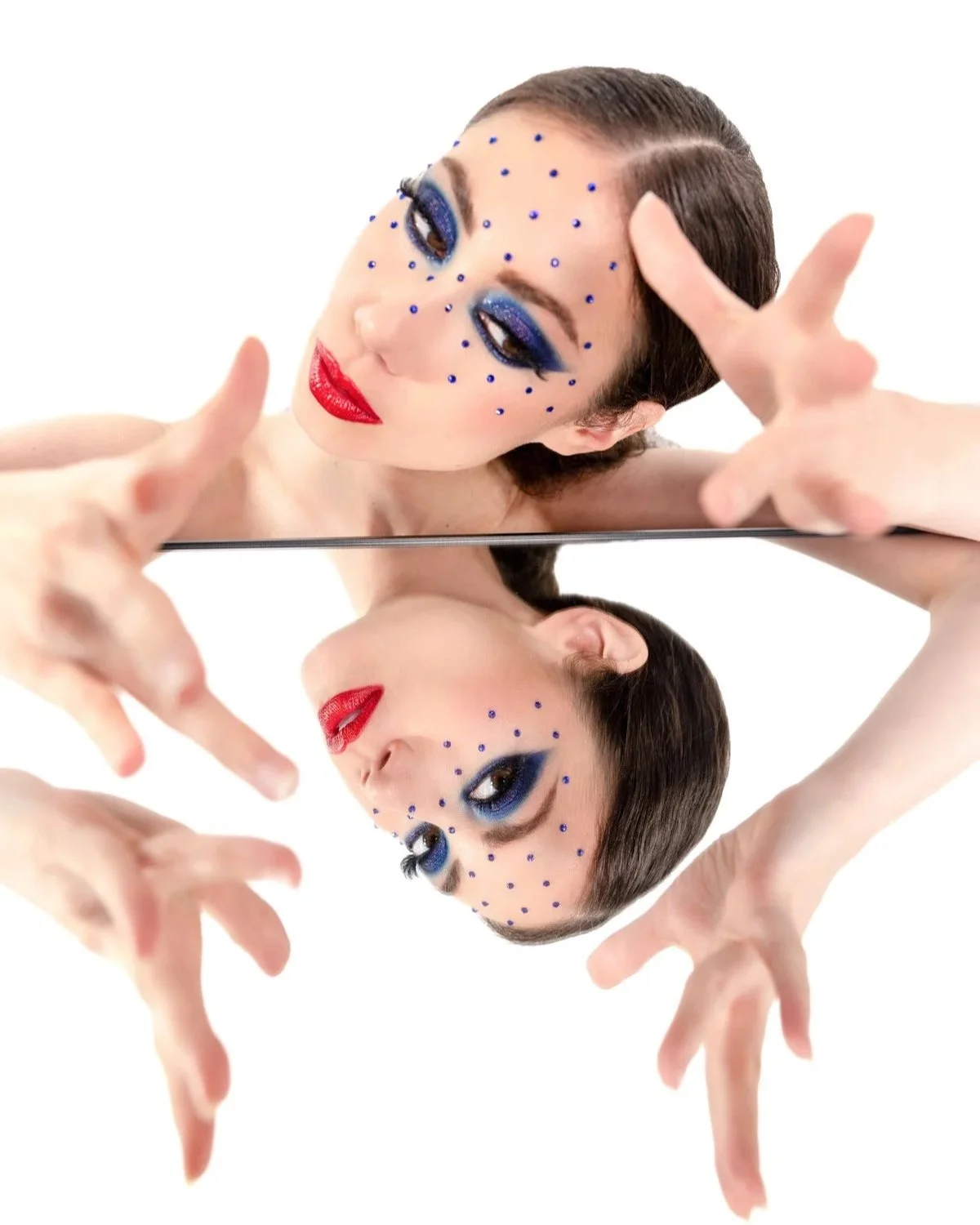Woman with creative makeup and blue rhinestones, reflected in a mirror, against a white background.