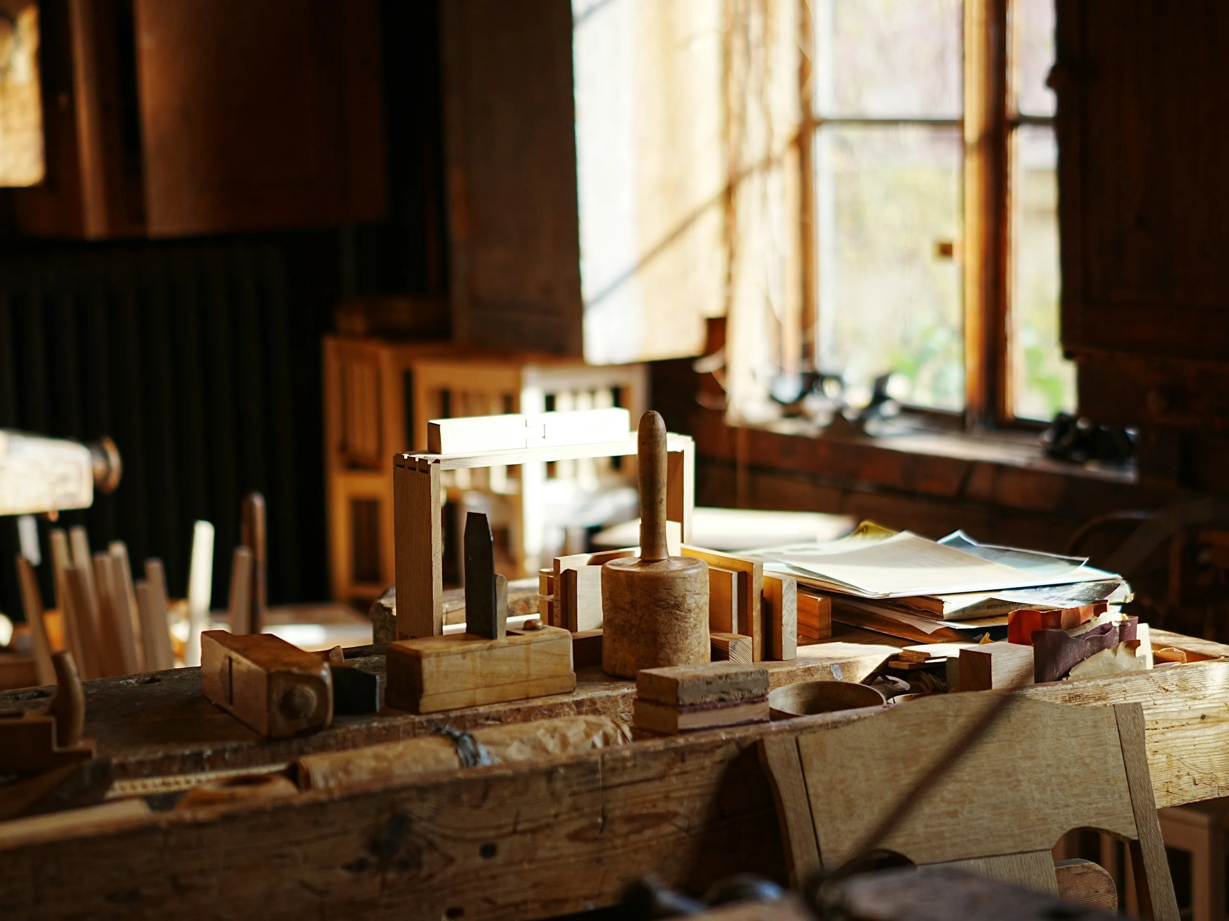 Workshop with woodworking tools and models on a wooden table, lit by natural light through windows.