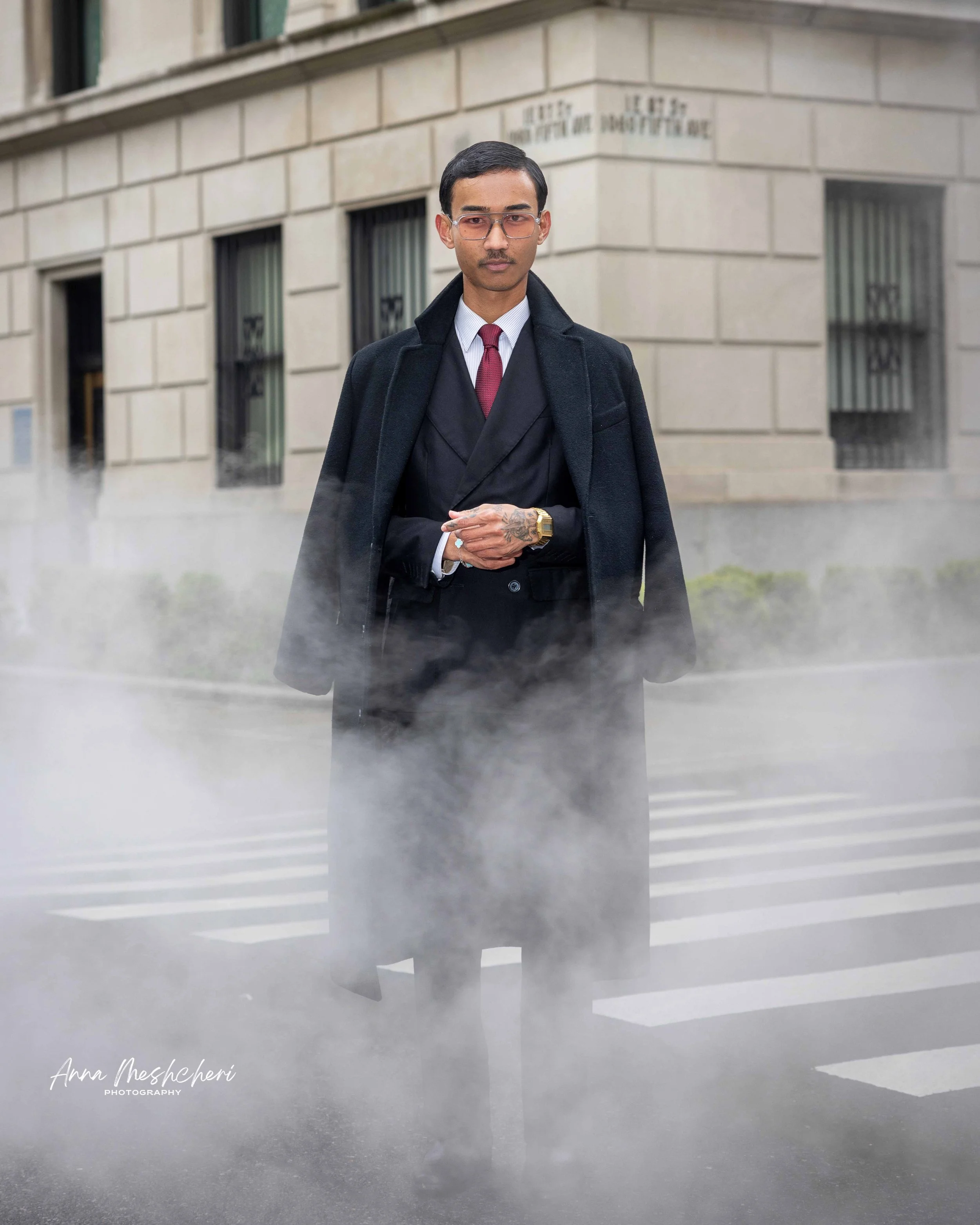 A man posing in a black suit and a black coat in a street of New York surrounded by smoke