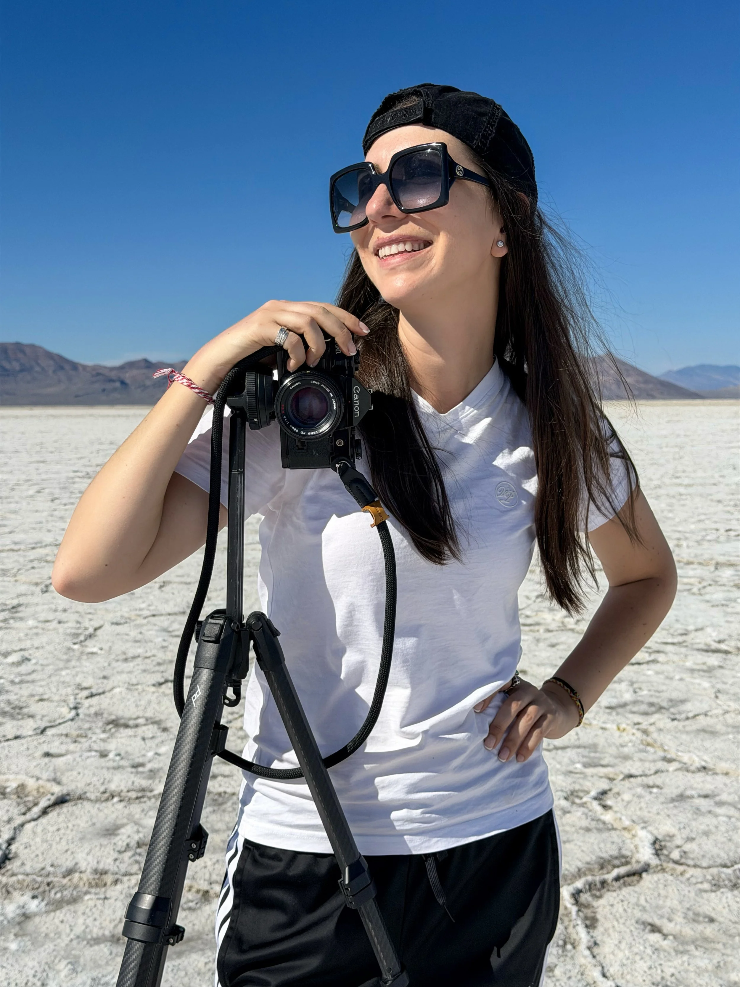 A woman with long dark hair, wearing large sunglasses, a backward black cap, a white T-shirt, and black shorts, stands outdoors on a salt flat or desert with mountains in the background, smiling while holding a camera on a tripod.
