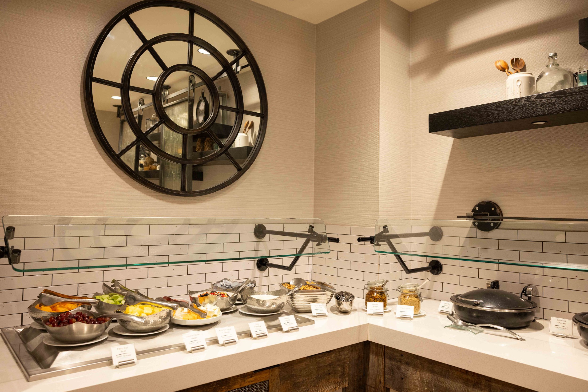 A buffet display with various bowls of fresh fruit, cereals, and toppings, set against a tiled wall with a large decorative mirror on the wall.