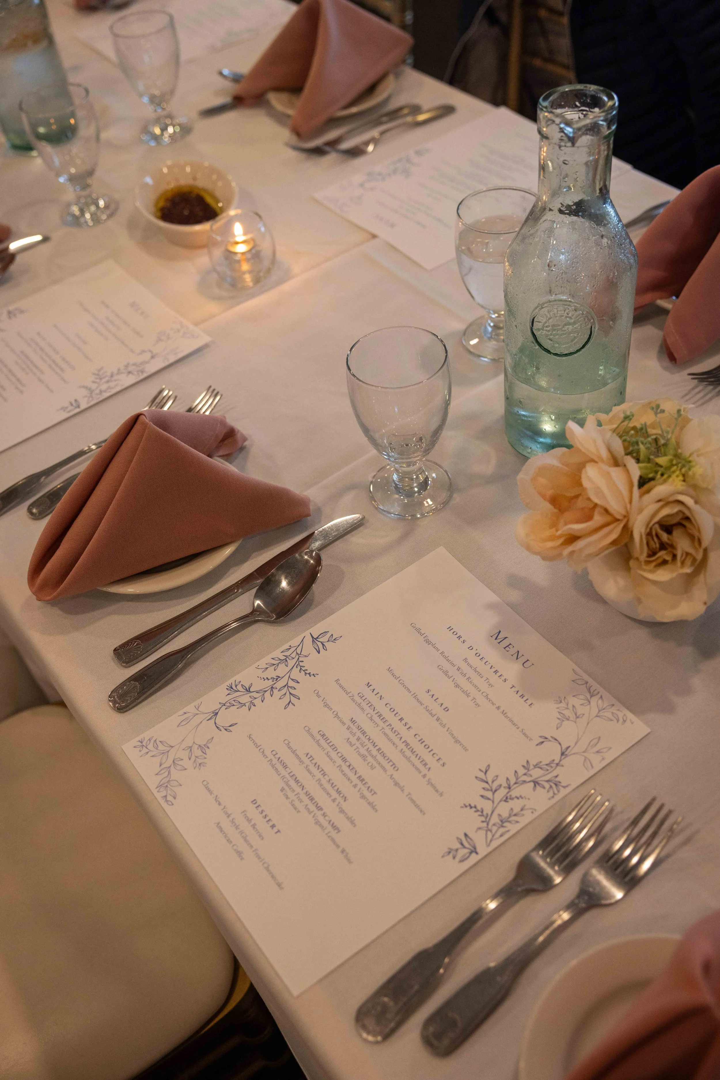 A table setting at a formal restaurant with a menu, pink cloth napkin, glass water pitcher, glasses for drinks, silverware, a small candle, a bowl of sauce, and a flower arrangement on a white tablecloth.
