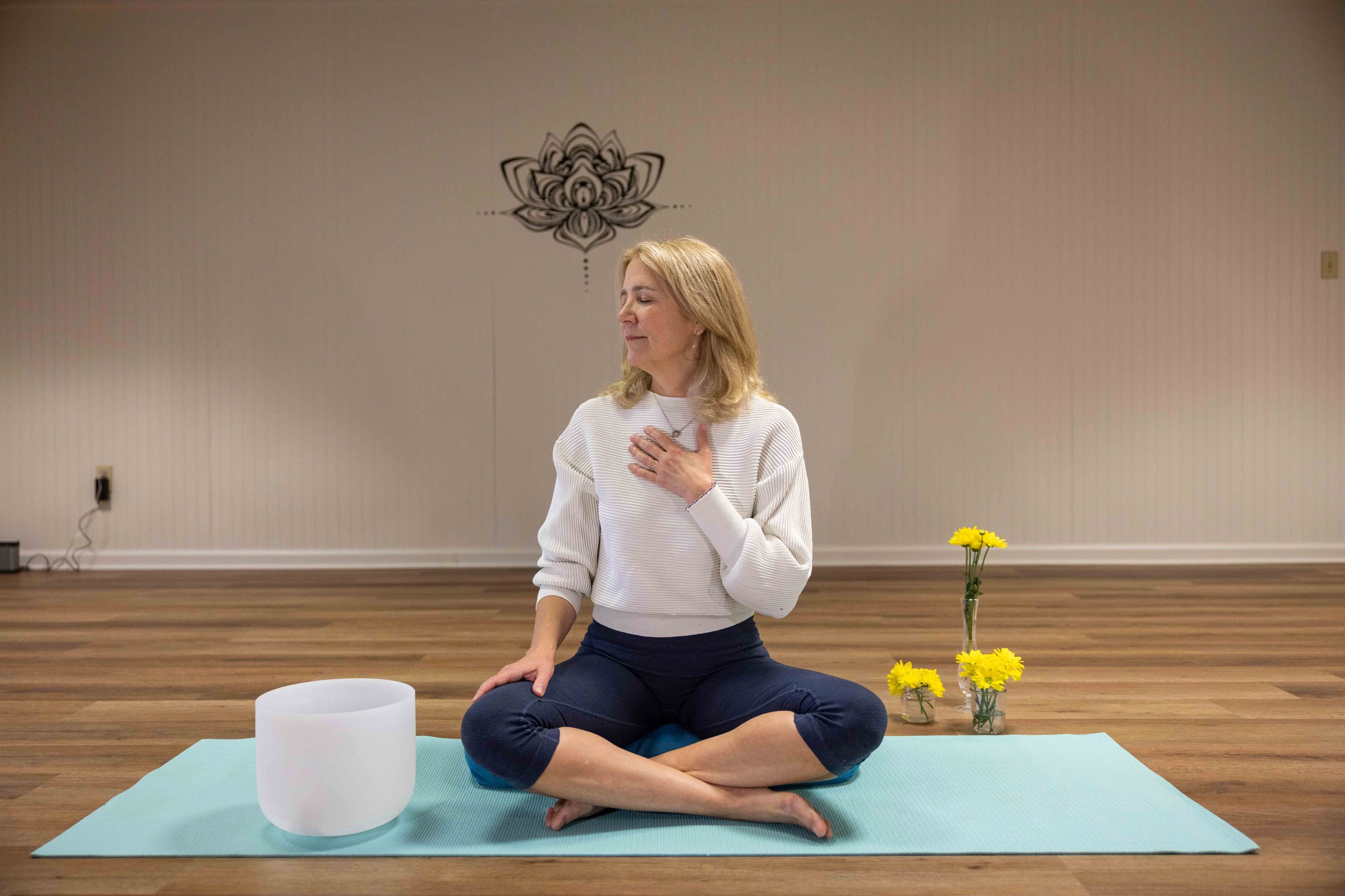 A woman practicing yoga indoors, sitting cross-legged on a mat with her hand over her heart. There is a singing bowl or similar object and yellow flowers on the floor near her. She has her eyes closed and appears peaceful.