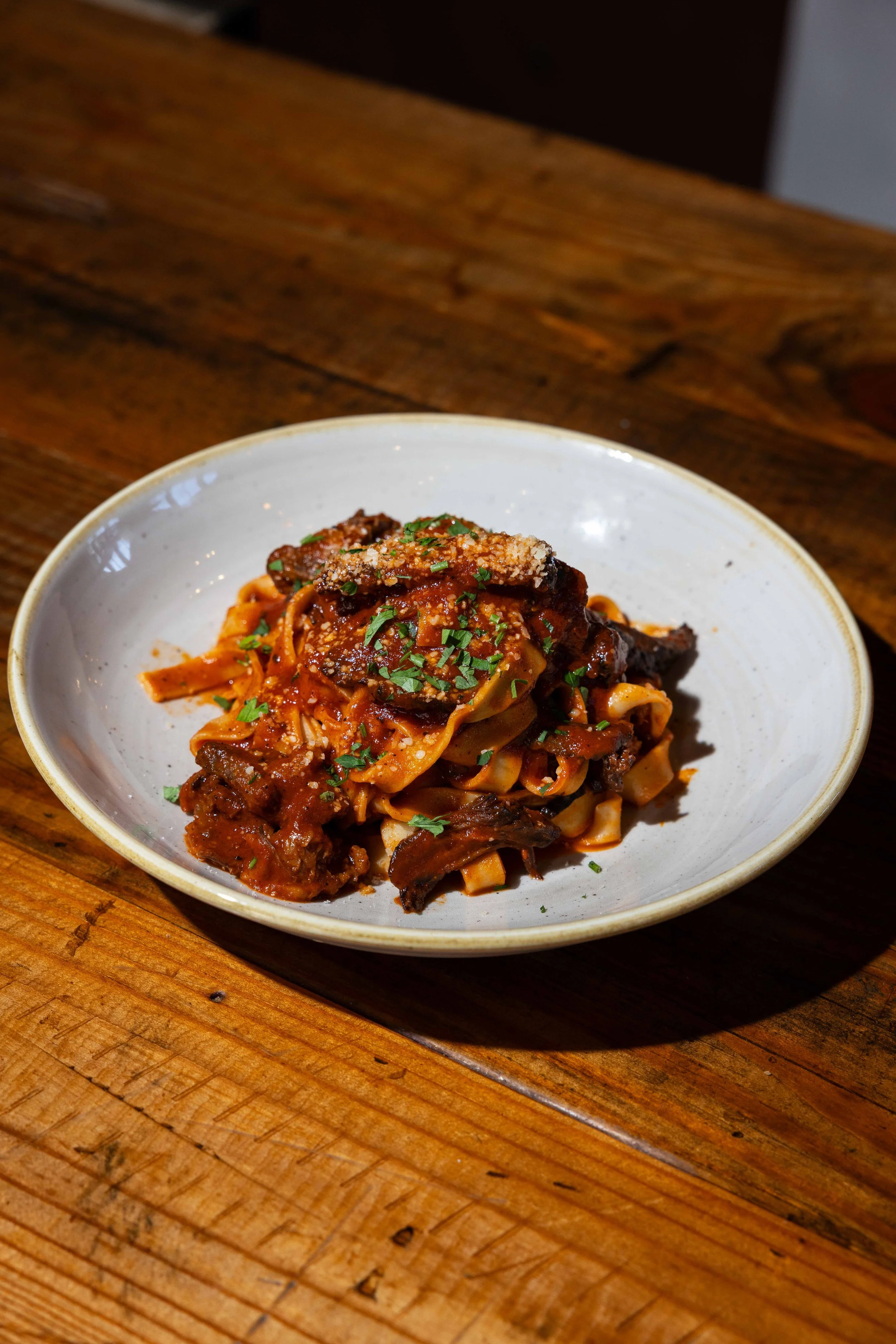 A bowl of pasta with beef, tomato sauce, garnished with chopped herbs and cheese, on a wooden table.