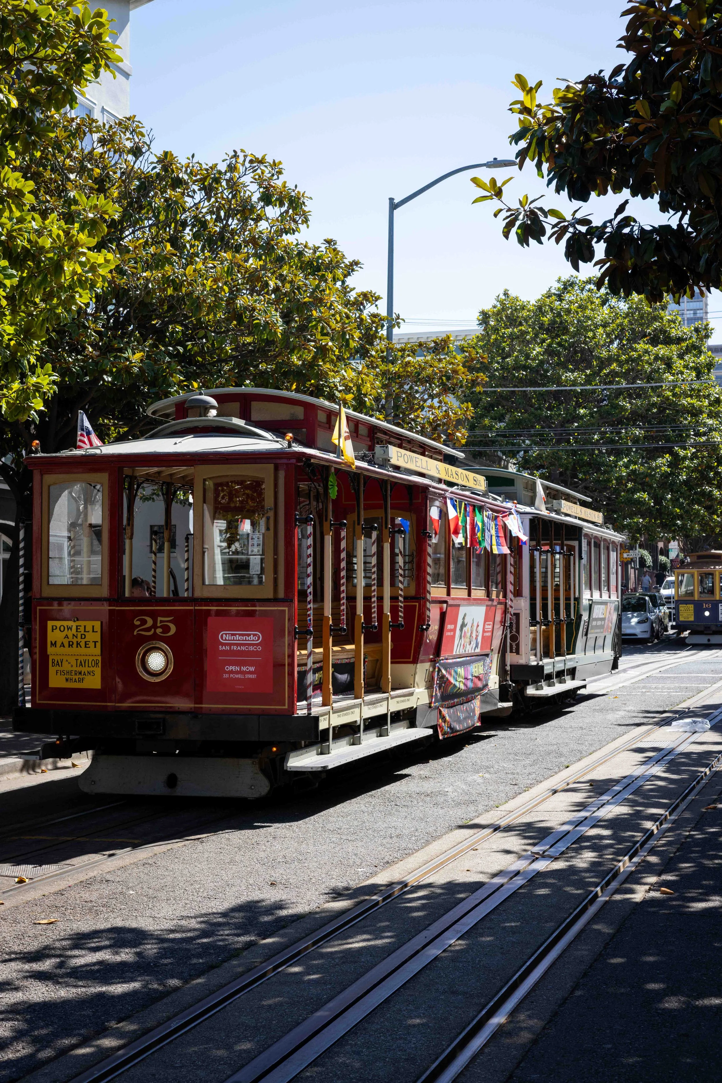Red trolley with advertisements for Powell and Market Street in San Francisco on a street with trees, including trolley tracks on the road, shadows of trees on the pavement, and metal poles for overhead wires.