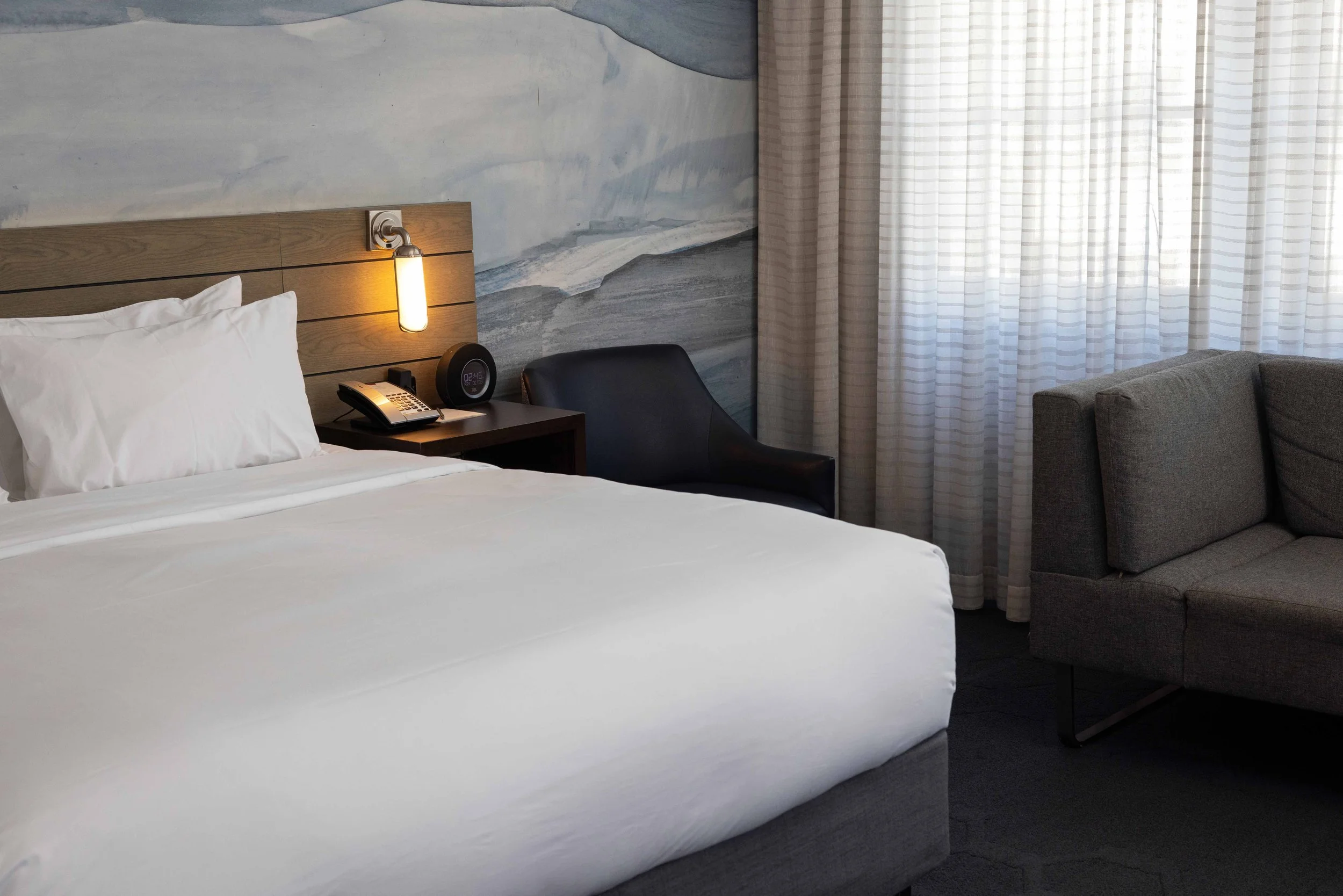 Hotel room with a bed, bedside table, clock, telephone, wall-mounted light, armchair, and curtains.