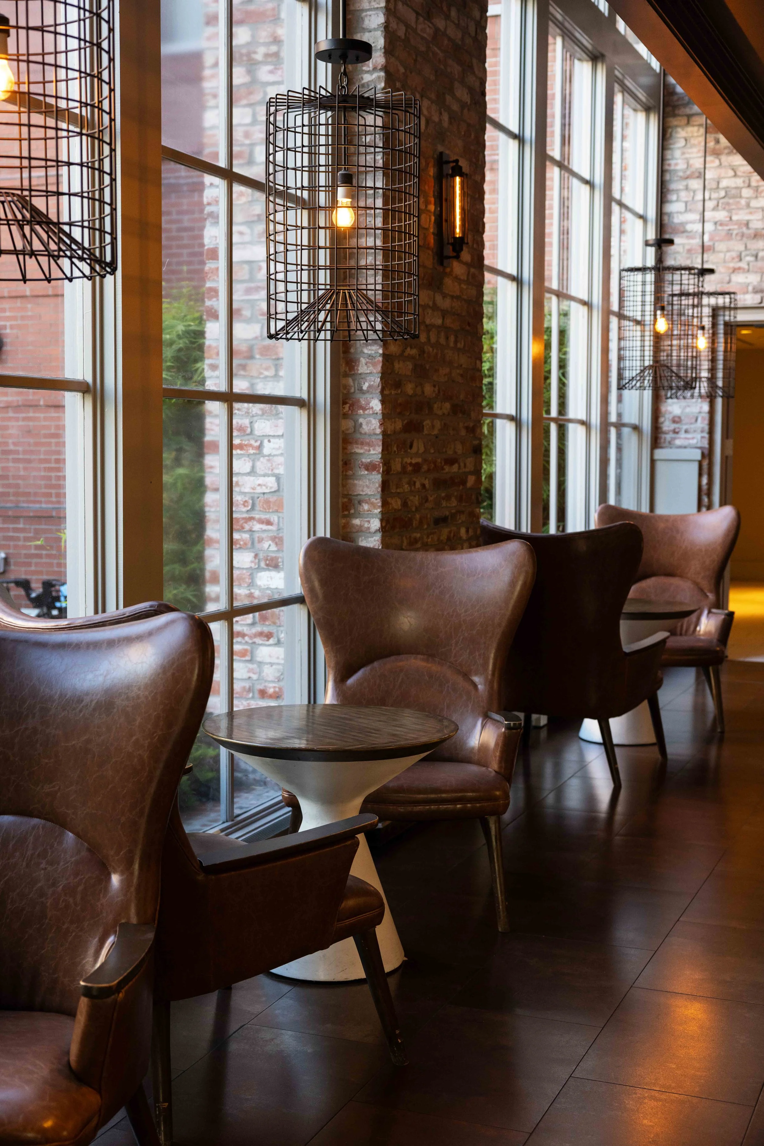 Interior of a modern cafe with leather chairs, large windows, and hanging wire-frame light fixtures, brick walls, and a small round table.