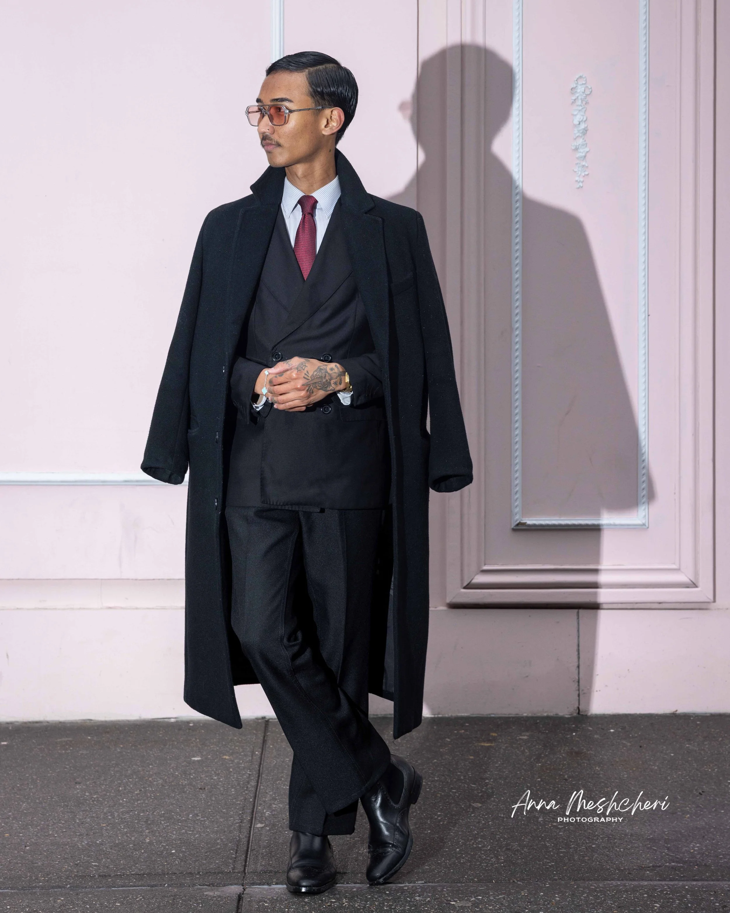A man wearing a suit and black coat standing in front of a pink wall
